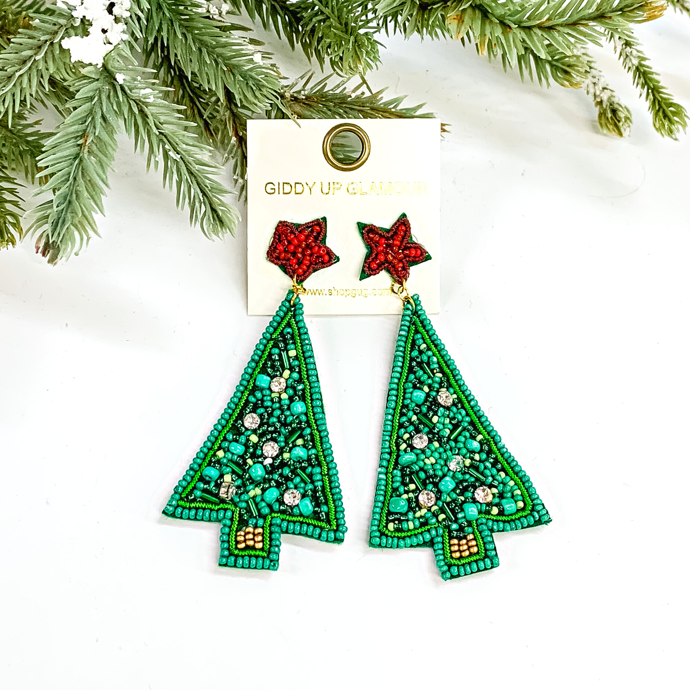 These are triangle shaped Christmas tree earrings in green with a red star  as the postback. There are different shades and shapes of green beads with  five clear crystals as ornaments. These earrings are taken on a white background with a tree in the back with snow as decor.