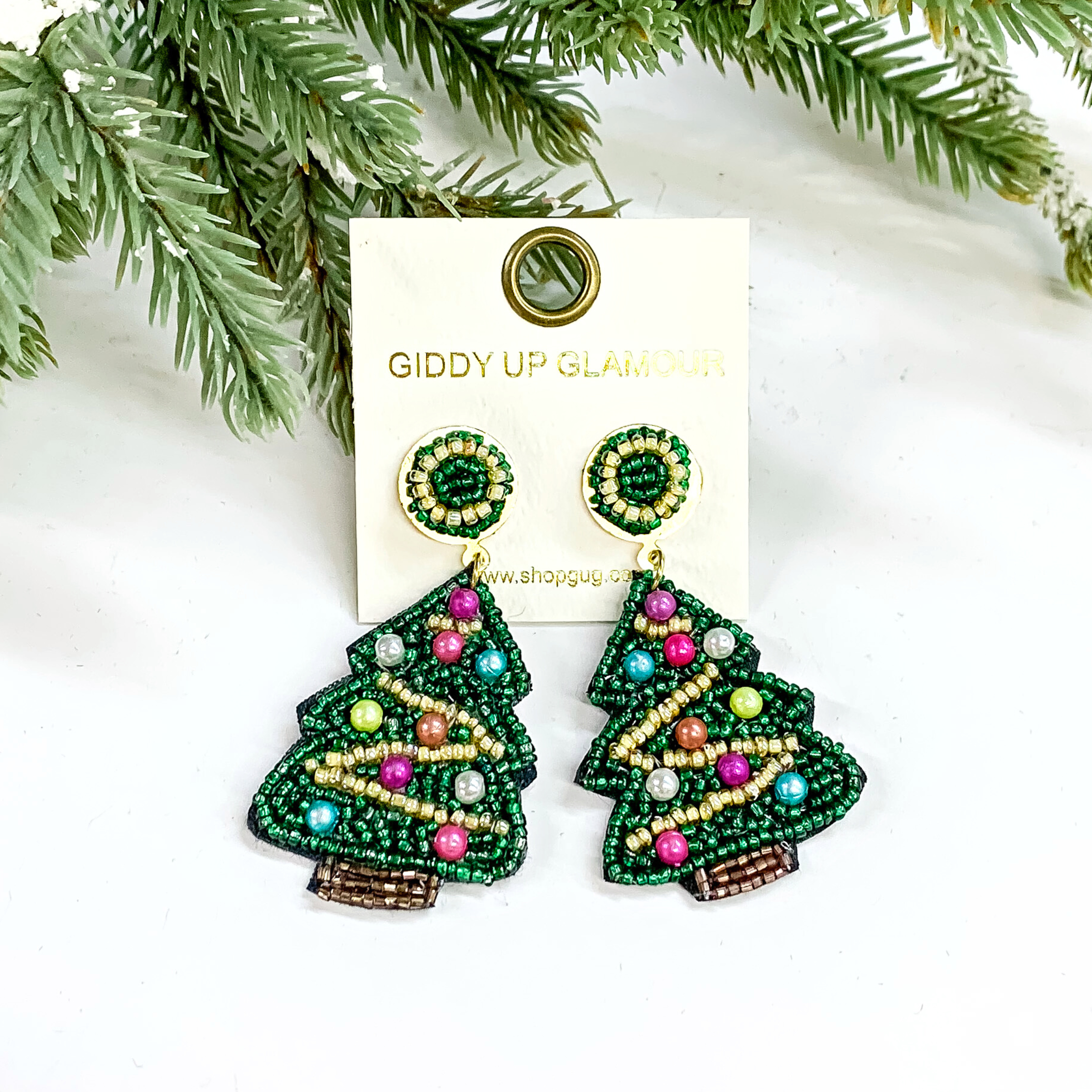 These are beaded Christmas tree earrings in green, gold, brown and multicolor pearl ornaments. There is champagne/gold beads as the 'ribbon' all around with the tree with pearl ornaments in purple,pink, teal silver, and rose gold. These earrings are taken on a white background with a tree in the back as decor.