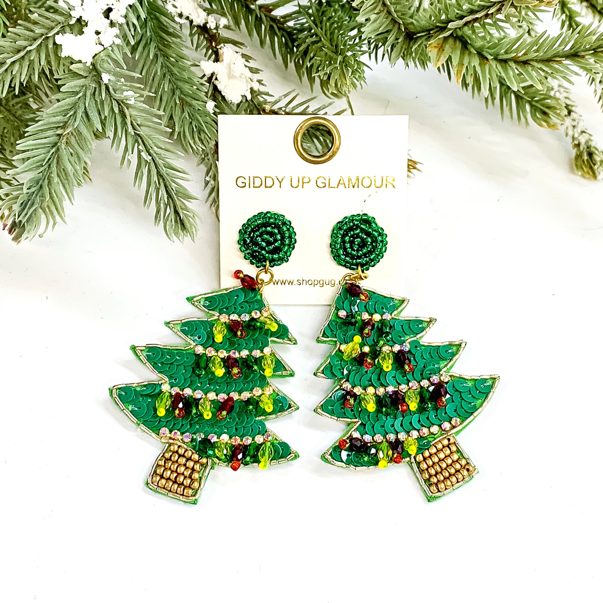 These are green Christmas tree earrings with beads and sequins in different colors. The tree earrings have green squins with a gold trim all around, the 'ribbon' are small ab crystals and the 'light' are small, teardrop crystals in red, green, and yellow. The stem has gold beads. These earrings are taken on a white background with a green tree and snow as decor.