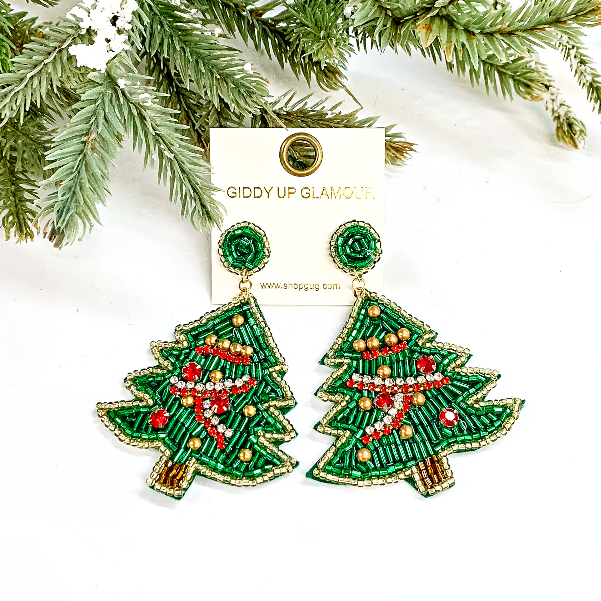 These are green beaded christmas tree earrings in different shapes and colors of beads. The tree has long green beads with a gold beaded outline and brown beaded stem. There are red and clear small crystals as the 'ribbon' , small red crystals and gold beads as oranaments. These earrings are taken on a white background with a green tree and snow in the back as decor.