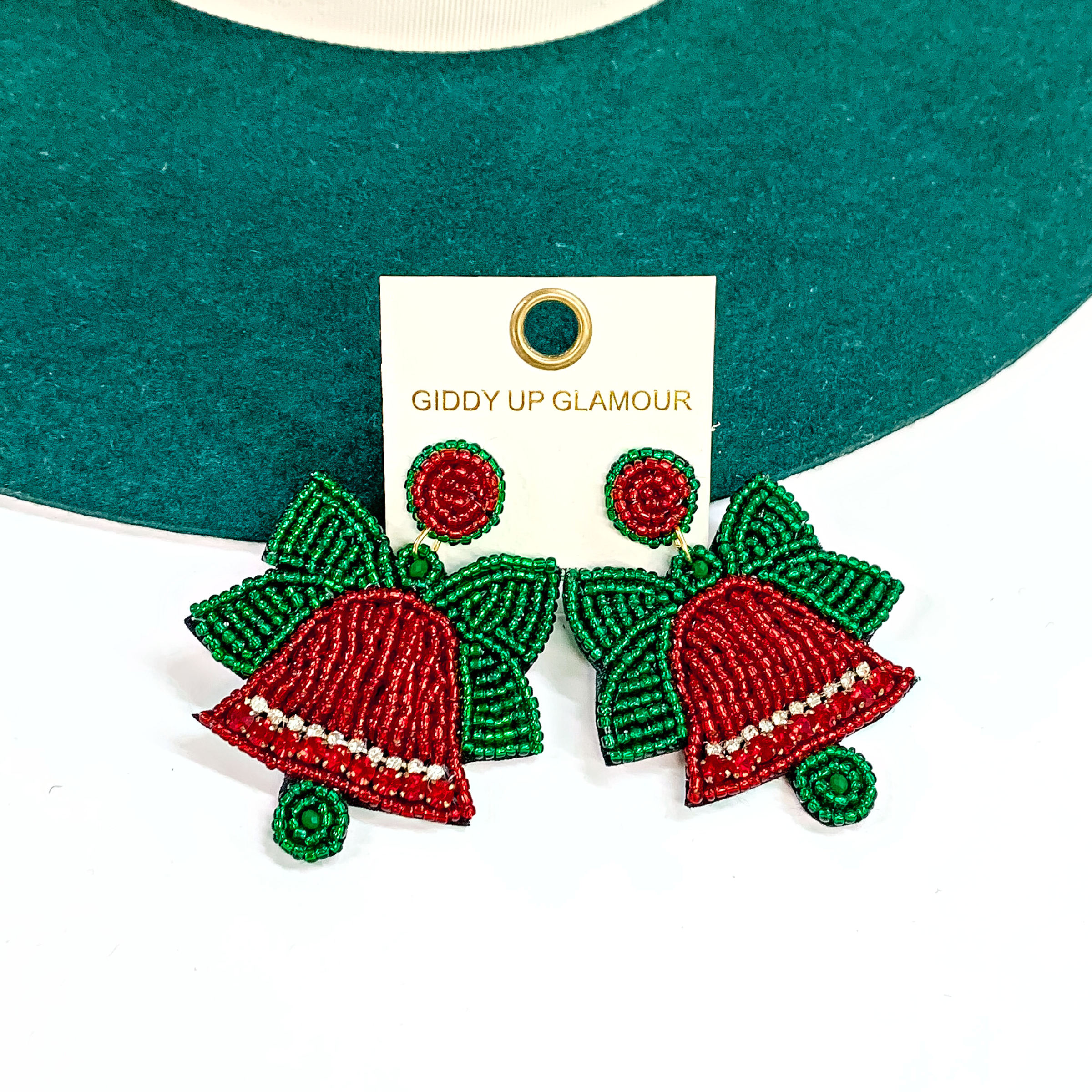 These are Christmas bell red and green seedbeaded earrings with a clear crystal bad across the red bell. The bell and postback are red, there is a green seedbeaded bwon on top of the bell. These earrings are taken laying on a dark green felt hat brim and on a white background.