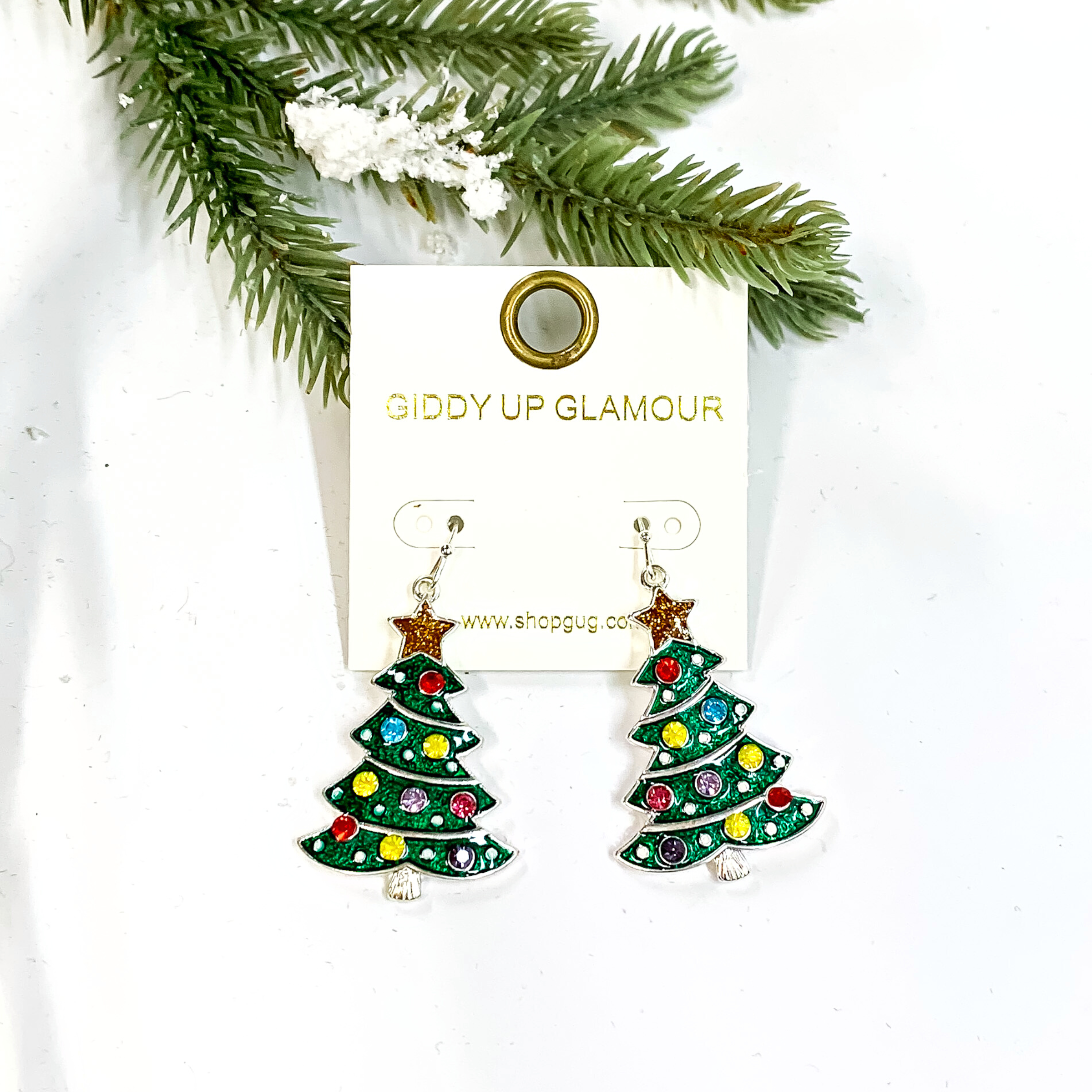 These are green Christmas tree earrings in a silver setting with a gold star on top. The tree has multicolor rhinetsones in red, light purple,purple, pink, yellow, and turquoise as 'ornaments'. These earrings are laying on a white background with a green tree  and snow in the back as decor,
