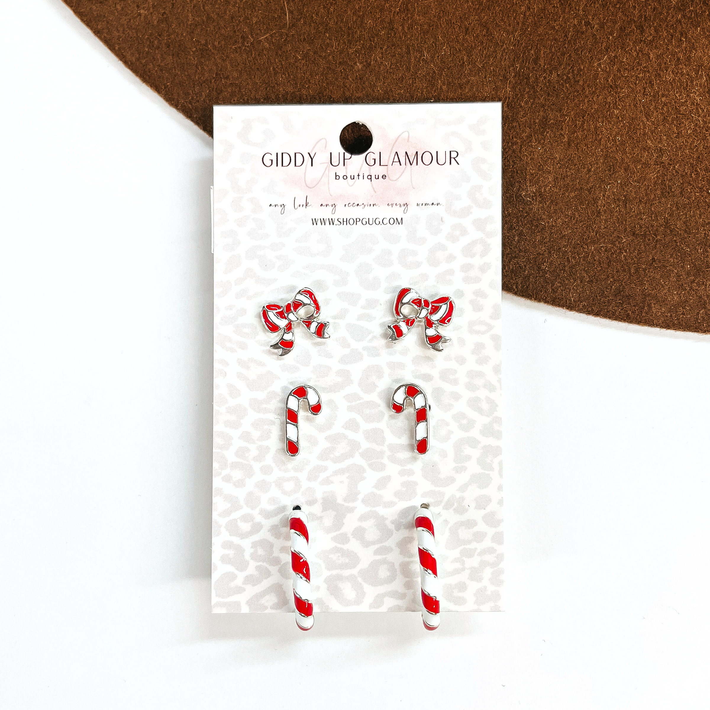 This is a set of three earrings in red and white in a silver setting. From top to bottom; bows, candy canes, and hoops. This earrings are placed in a Giddy Up Glamour card holder. This earring set is taken on a white background and on a dark brown felt hat brim.