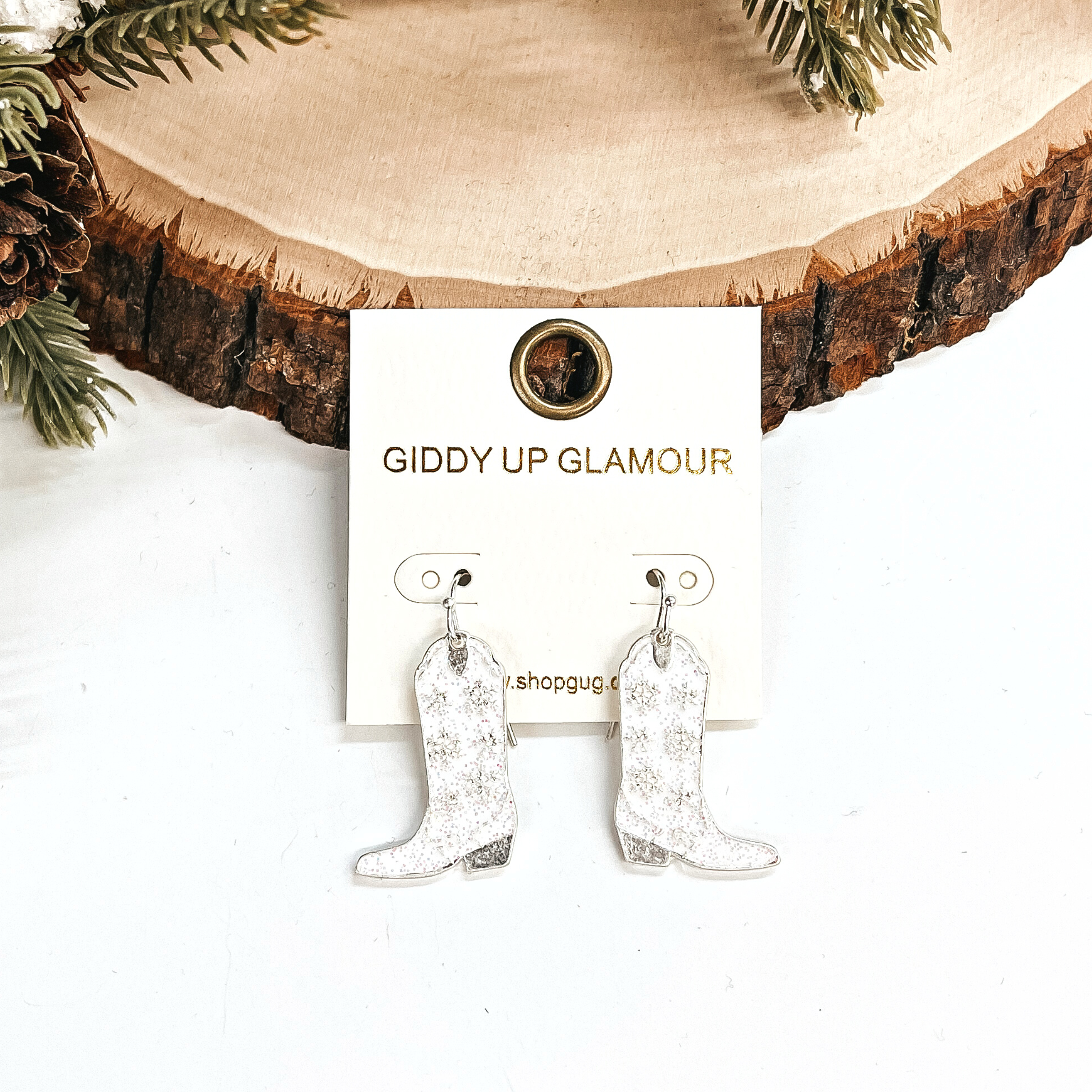 These are boot pendant drop earrings in white glitter and silver.  The earrings have a snowflake pattern in silver. These earrings are taken  leaning up against a slab of wood and on a white background with a pine cone  tree in the back as decor.