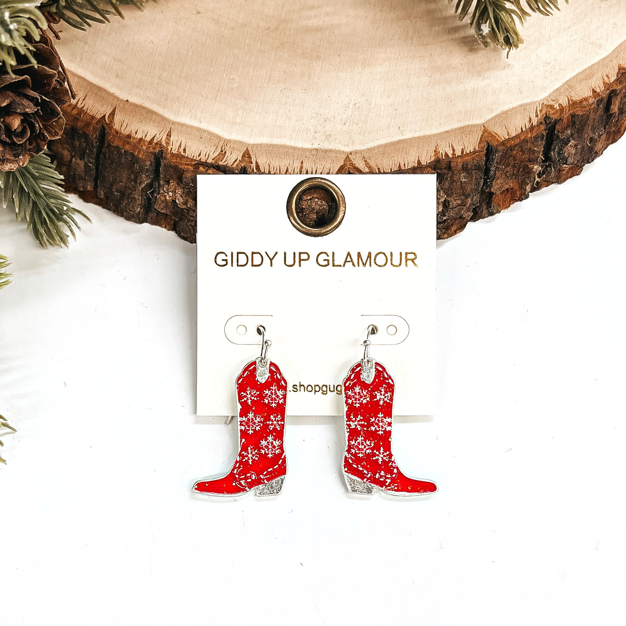 These are boot pendant drop earrings in red glitter and silver.  The earrings have a snowflake pattern in silver. These earrings are taken  leaning up against a slab of wood and on a white background with a pine cone  tree in the back as decor.