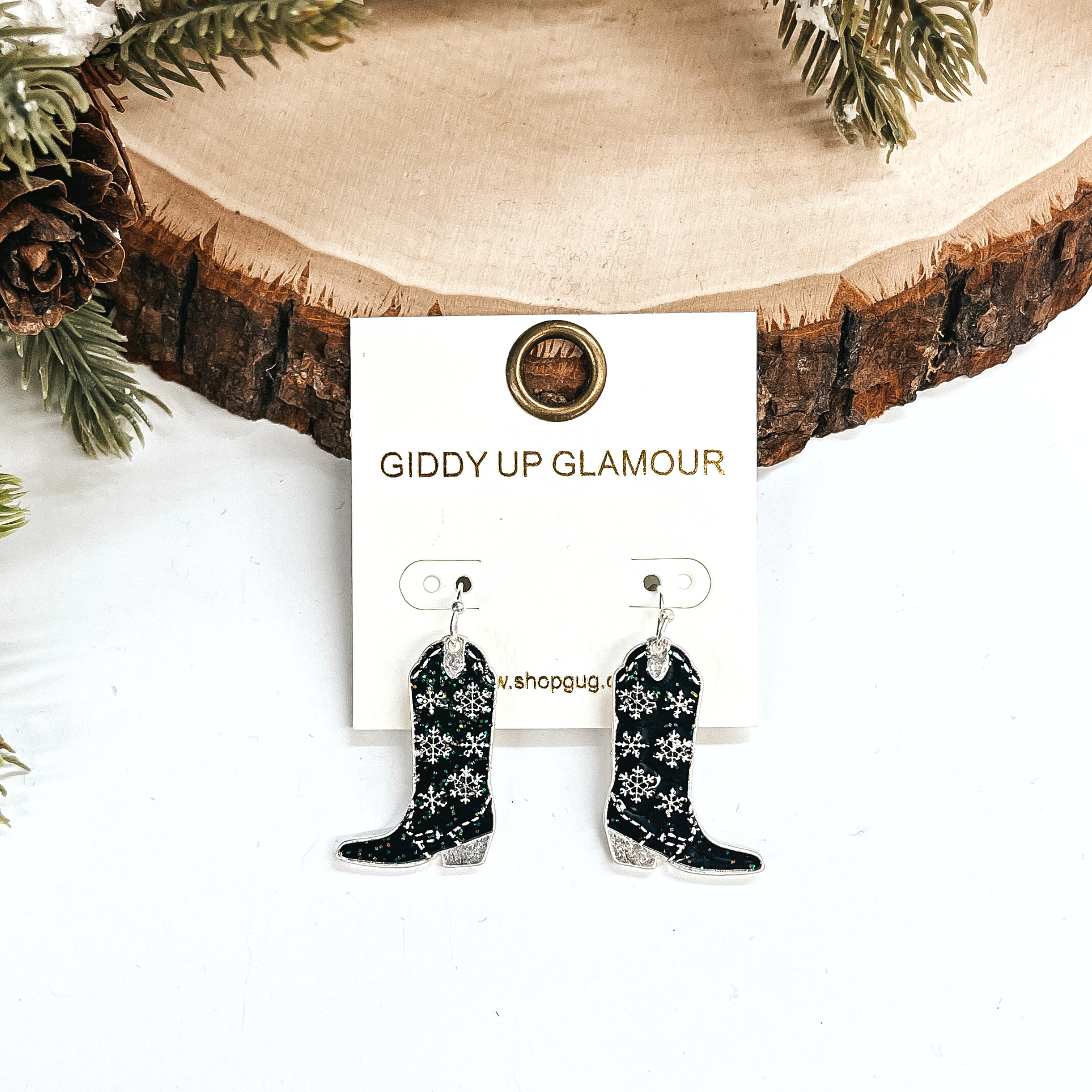 These are boot pendant drop earrings in black glitter and silver.  The earrings have a snowflake pattern in silver. These earrings are taken  leaning up against a slab of wood and on a white background with a pine cone  tree in the back as decor.