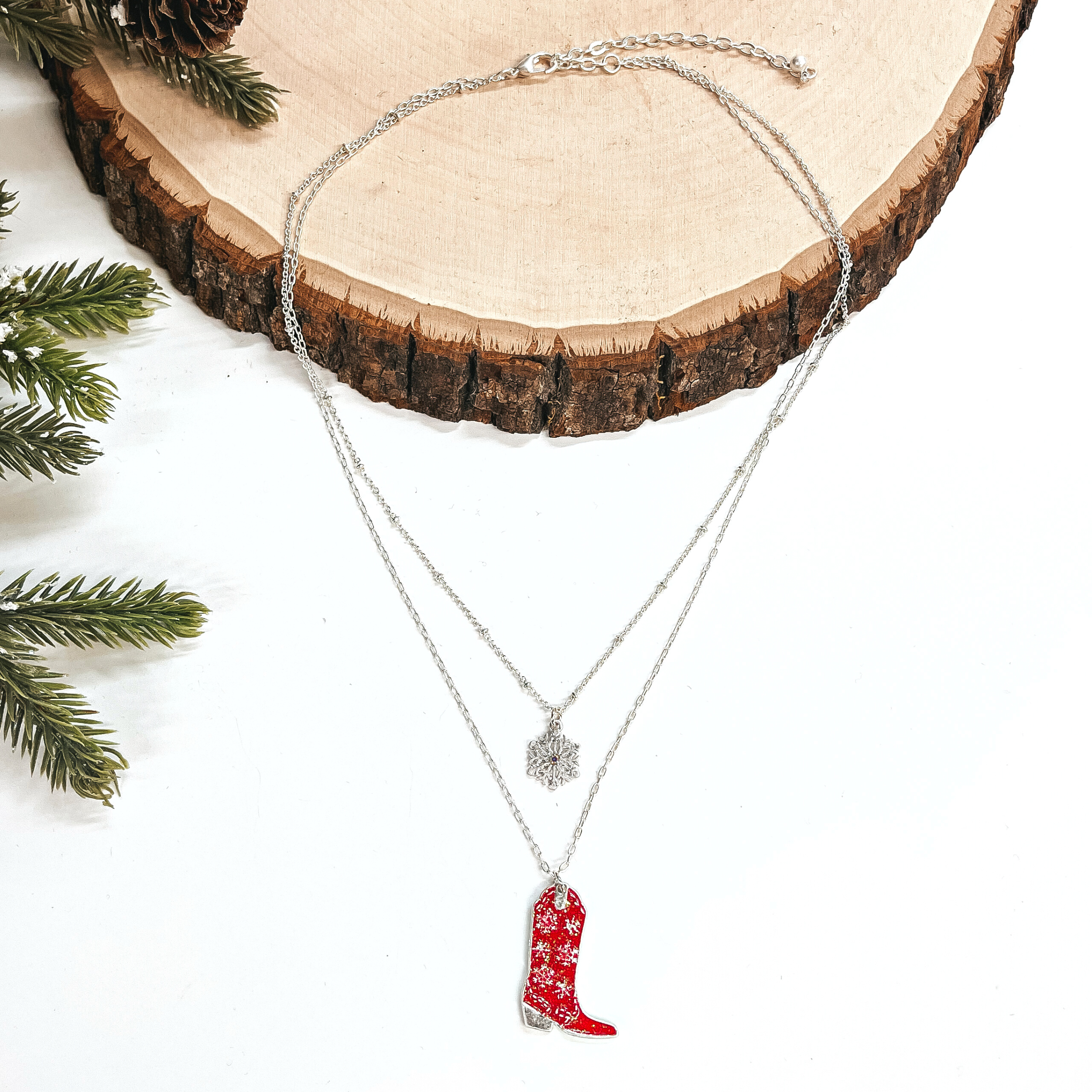 This is a double strand silver necklace with two hanging pendants.  The smaller strand has a silver snowflake charm and the longer strand has a  red glitter boot pendant. The boot pendant has a silver snowflake pattern  and the boot pendant is in a silver setting. This necklace is laying on a  white background and a slab of wood, with a pine cone tree in the side as decor.