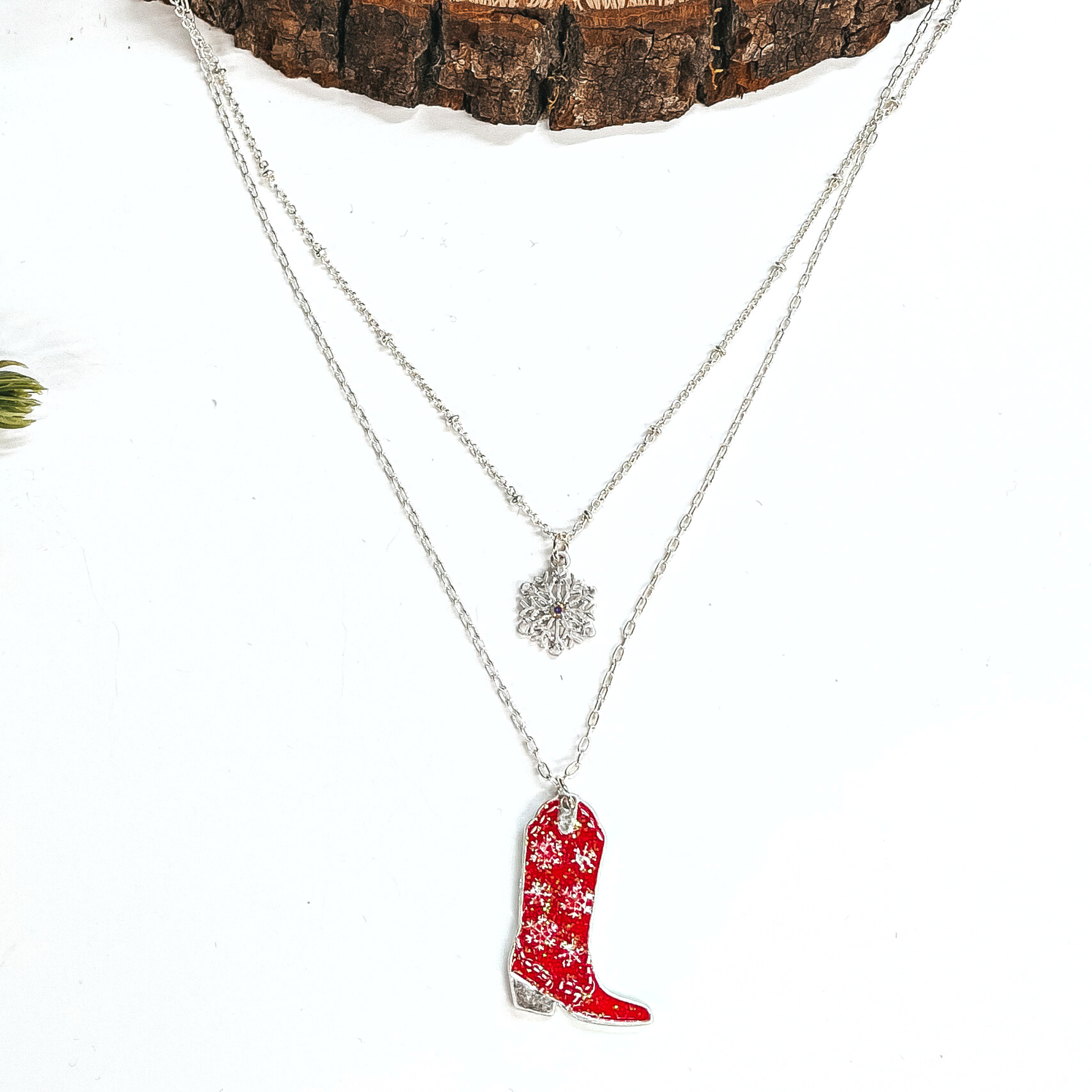 This is a double strand silver necklace with two hanging pendants.  The smaller strand has a silver snowflake charm and the longer strand has a  red glitter boot pendant. The boot pendant has a silver snowflake pattern  and the boot pendant is in a silver setting. This necklace is laying on a  white background and a slab of wood, with a pine cone tree in the side as decor.