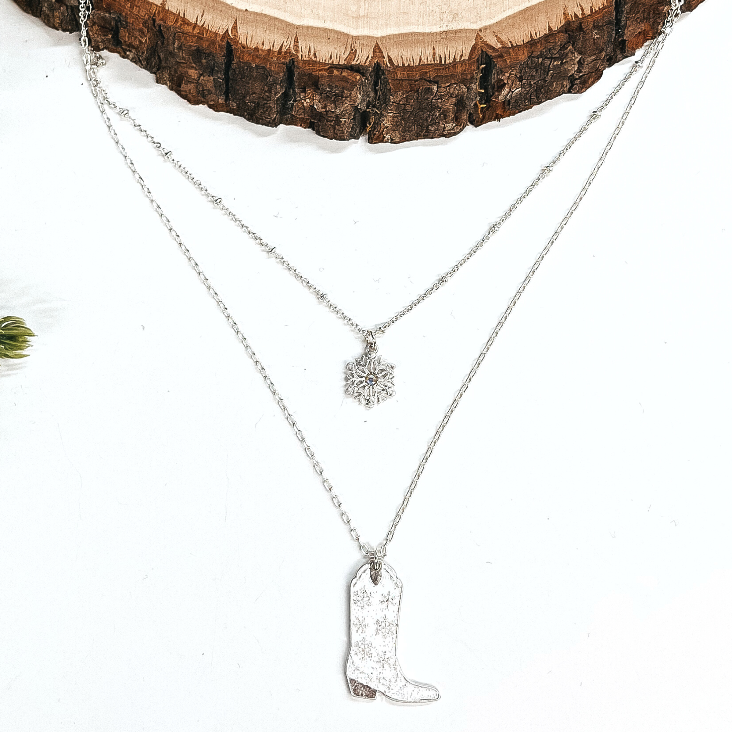 This is a double strand silver necklace with two hanging pendants.  The smaller strand has a silver snowflake charm and the longer strand has a  white glitter boot pendant. The boot pendant has a silver snowflake pattern  and the boot pendant is in a silver setting. This necklace is laying on a  white background and a slab of wood, with a pine cone tree in the side as decor.