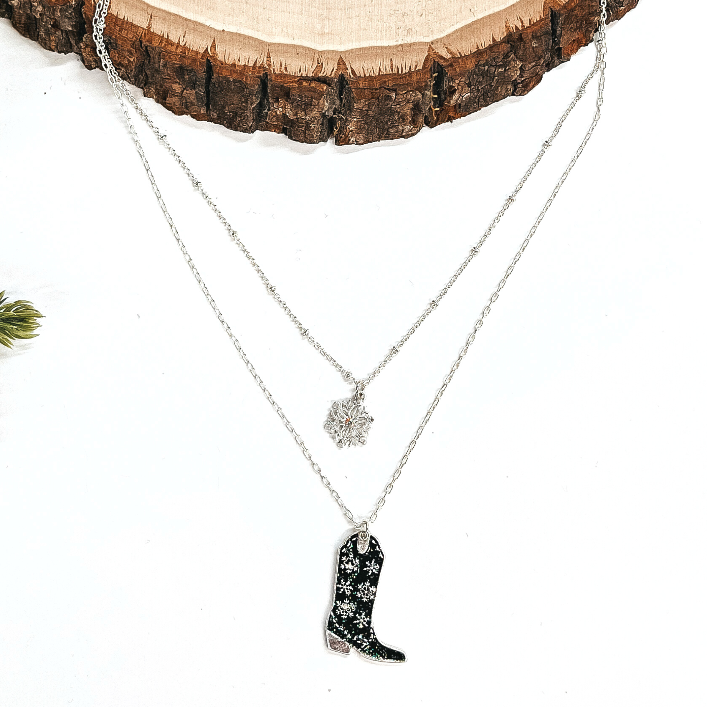 This is a double strand silver necklace with two hanging pendants.  The smaller strand has a silver snowflake charm and the longer strand has a  black glitter boot pendant. The boot pendant has a silver snowflake pattern  and the boot pendant is in a silver setting. This necklace is laying on a  white background and a slab of wood, with a pine cone tree in the side as decor.