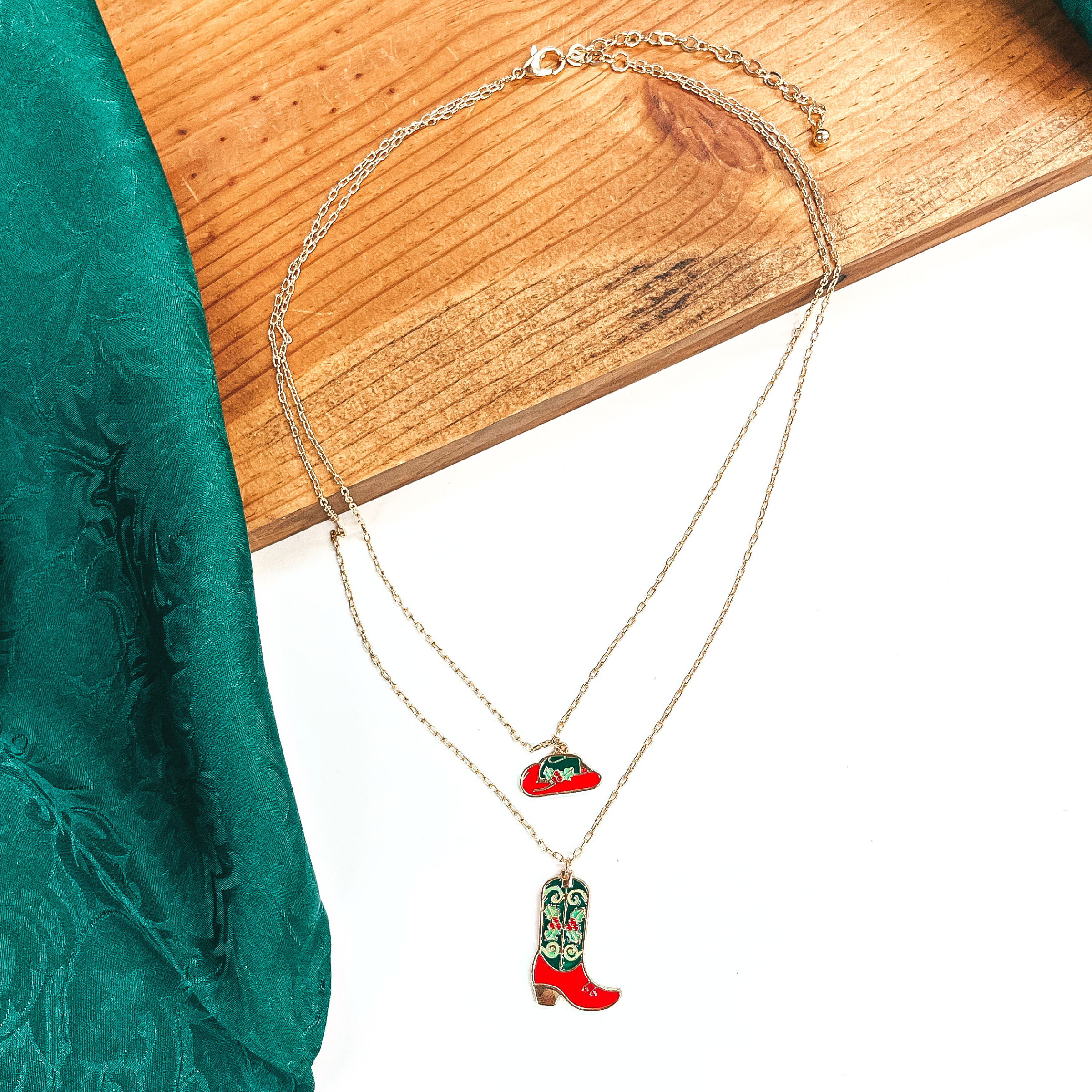 This is a double strand gold necklace with two hanging pendants.  The smaller strand has a hat pendant and the longer strand has a  boot pendant. Both pendants are red and dark green with mistletoe design  on them in a gold setting. This necklace is laying on a  white background and a slab of wood, with a green wild rag in the side as decor.