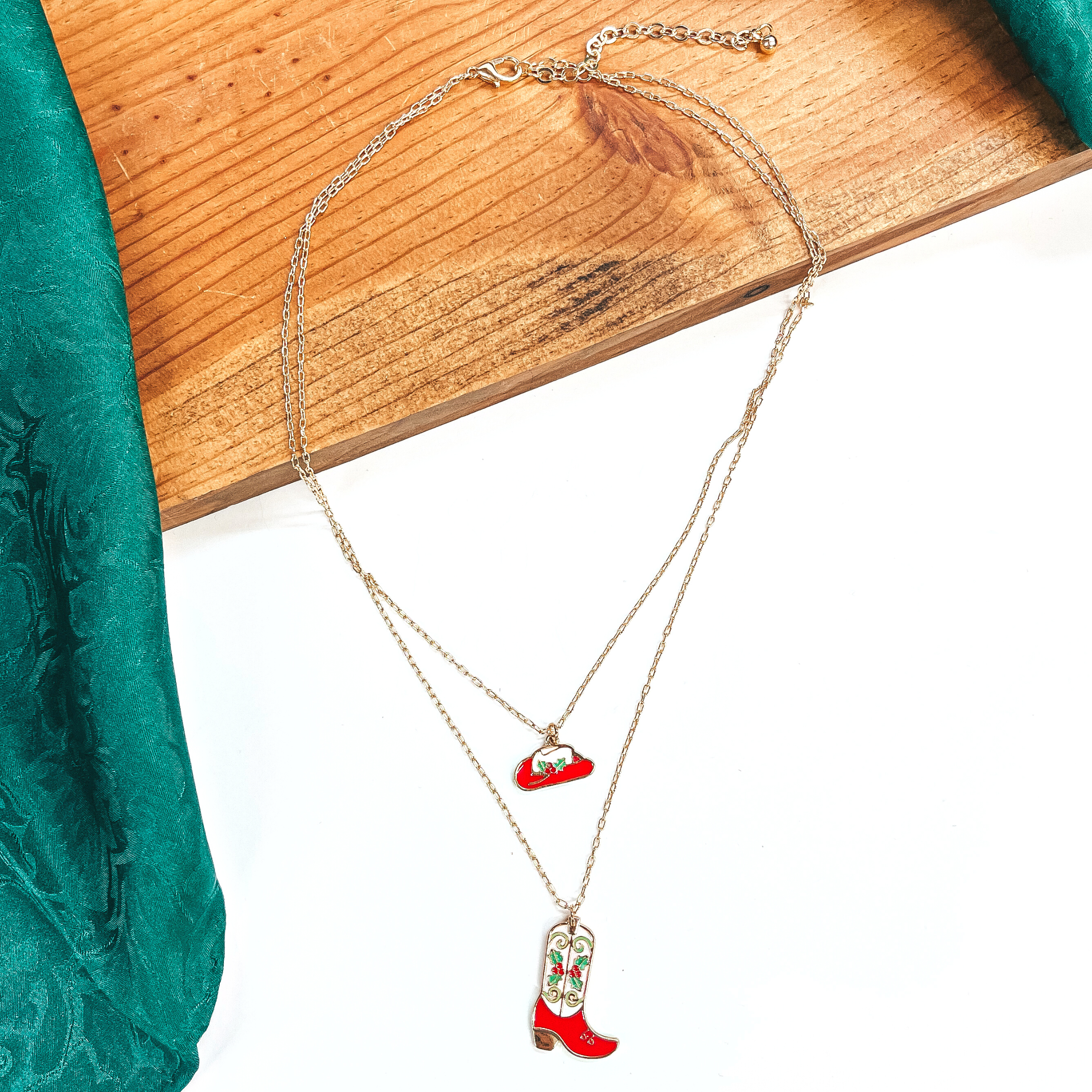This is a double strand gold necklace with two hanging pendants.  The smaller strand has a hat pendant and the longer strand has a  boot pendant. Both pendants are red and white with mistletoe design  on them in a gold setting. This necklace is laying on a  white background and a slab of wood, with a green wild rag in the side as decor.