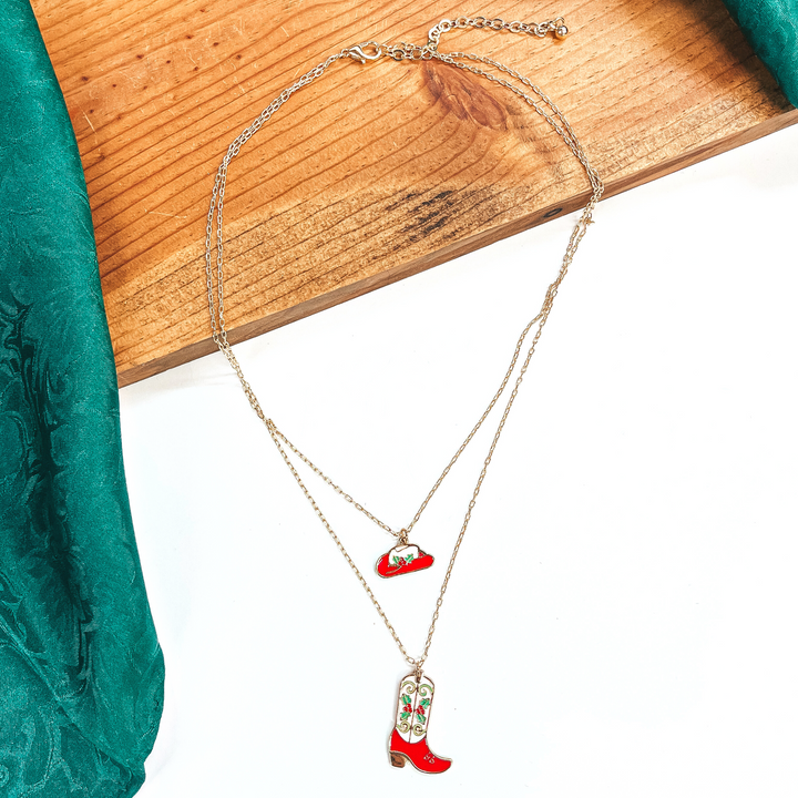 This is a double strand gold necklace with two hanging pendants.  The smaller strand has a hat pendant and the longer strand has a  boot pendant. Both pendants are red and white with mistletoe design  on them in a gold setting. This necklace is laying on a  white background and a slab of wood, with a green wild rag in the side as decor.