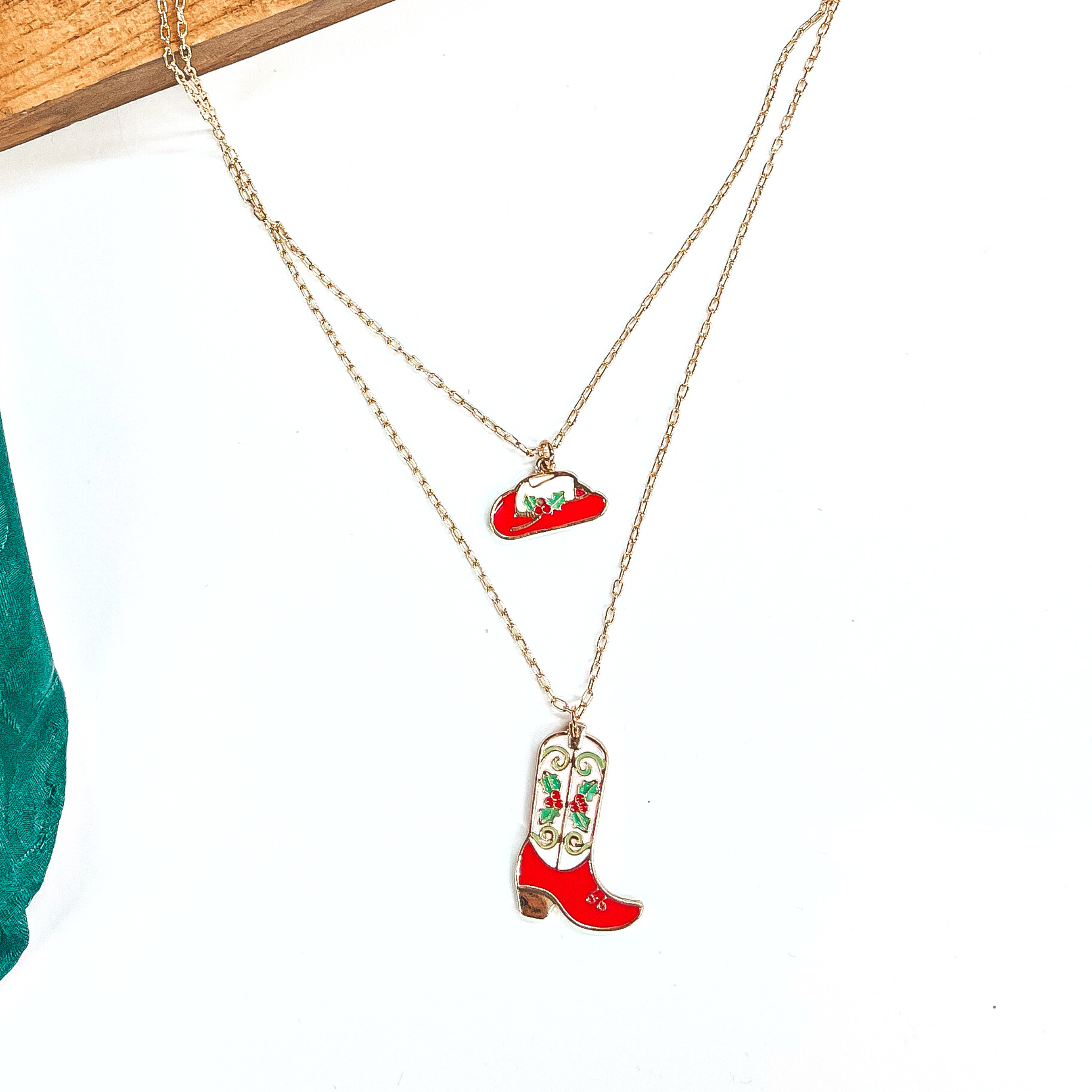 This is a double strand gold necklace with two hanging pendants.  The smaller strand has a hat pendant and the longer strand has a  boot pendant. Both pendants are red and white with mistletoe design  on them in a gold setting. This necklace is laying on a  white background and a slab of wood, with a green wild rag in the side as decor.