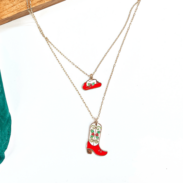 This is a double strand gold necklace with two hanging pendants.  The smaller strand has a hat pendant and the longer strand has a  boot pendant. Both pendants are red and white with mistletoe design  on them in a gold setting. This necklace is laying on a  white background and a slab of wood, with a green wild rag in the side as decor.