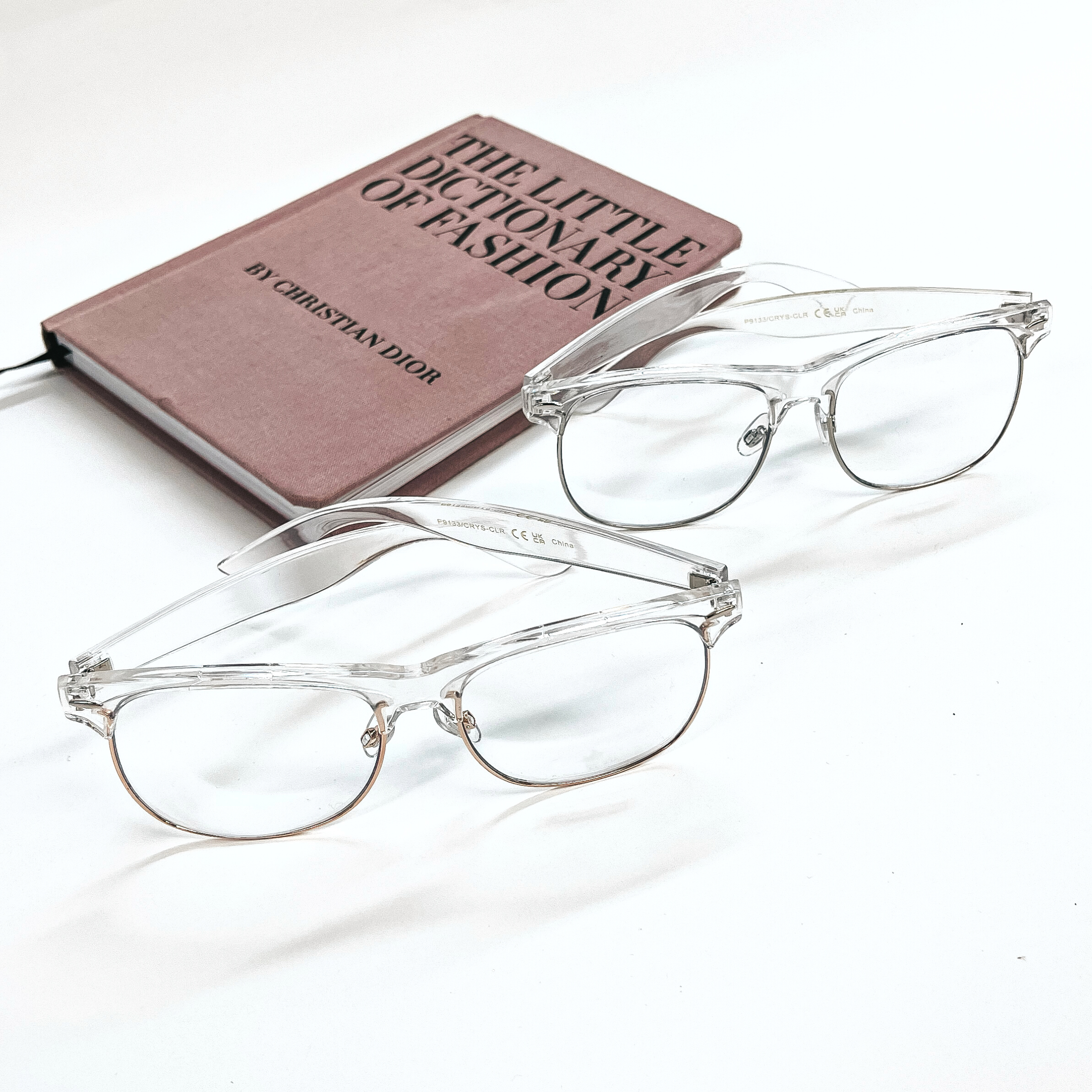 These are two pairs of clear glasses, both clear lenses and clear frames. The frames in front have a rose gold outline and the ones in the back have a silver outline. Both of these glasses are taken on a white background with a pink/purple fashion book in the back as decor.