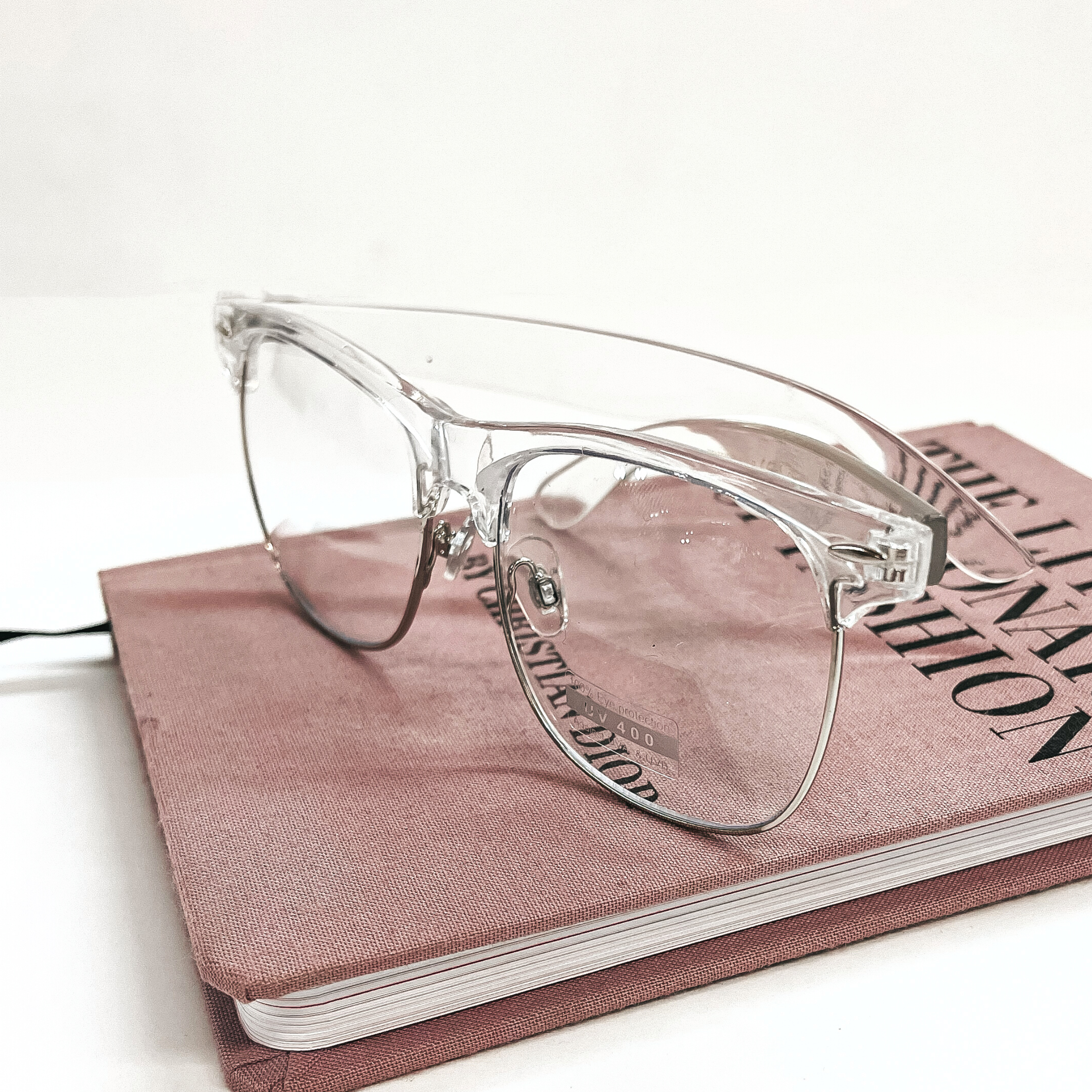 These are clear frame glasses with a clear lenses and a silver outline in the bottom. These glasses are taken on top of a pink/purple fashion book and on a white background.