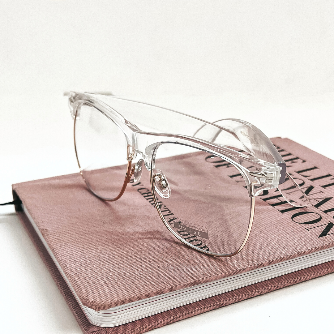 These are clear frame glasses with clear lenses and a rose gold outline in the bottom. These glasses are taken on a pink/purple fashion book and on a white background.