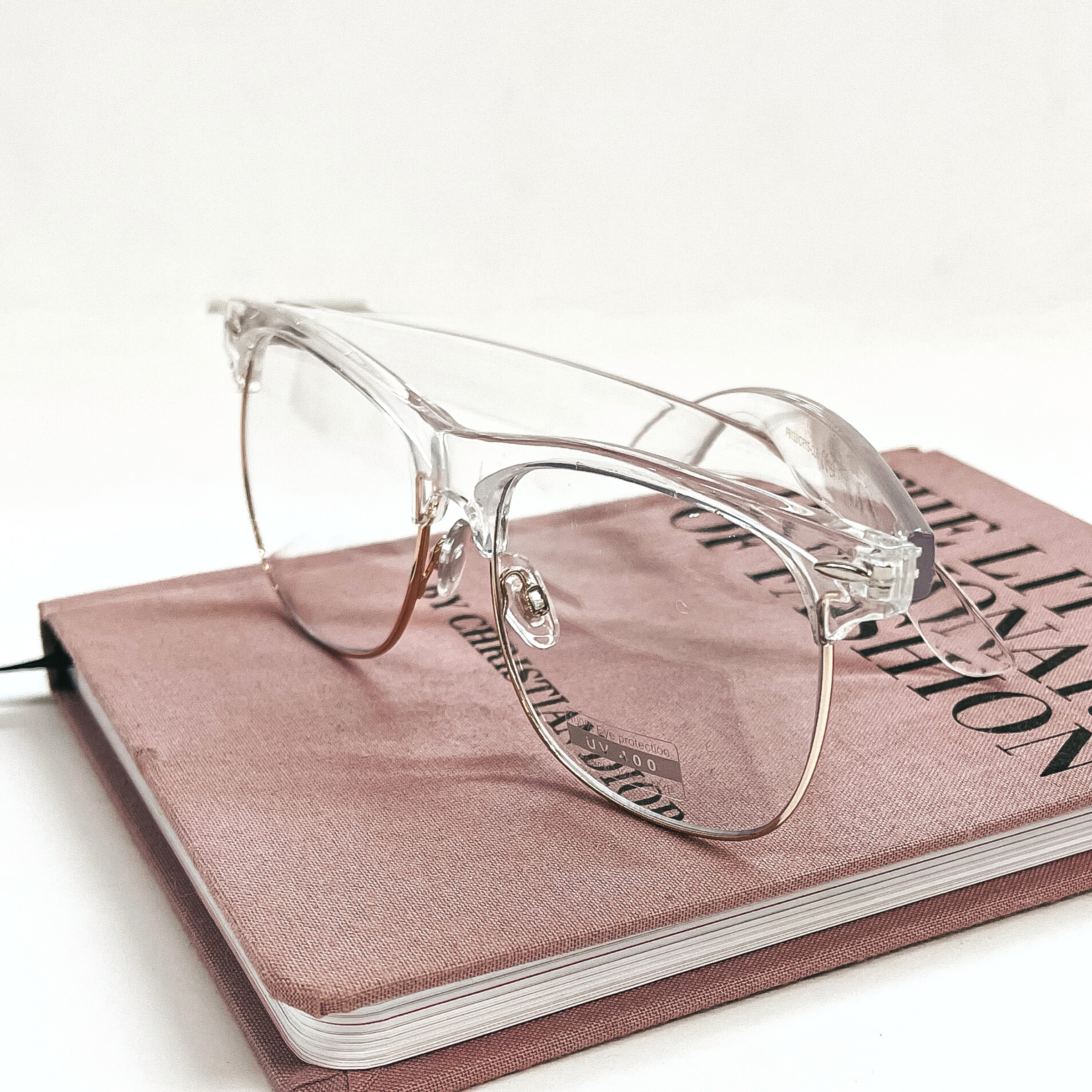 These are clear frame glasses with clear lenses and a rose gold outline in the bottom. These glasses are taken on a pink/purple fashion book and on a white background.