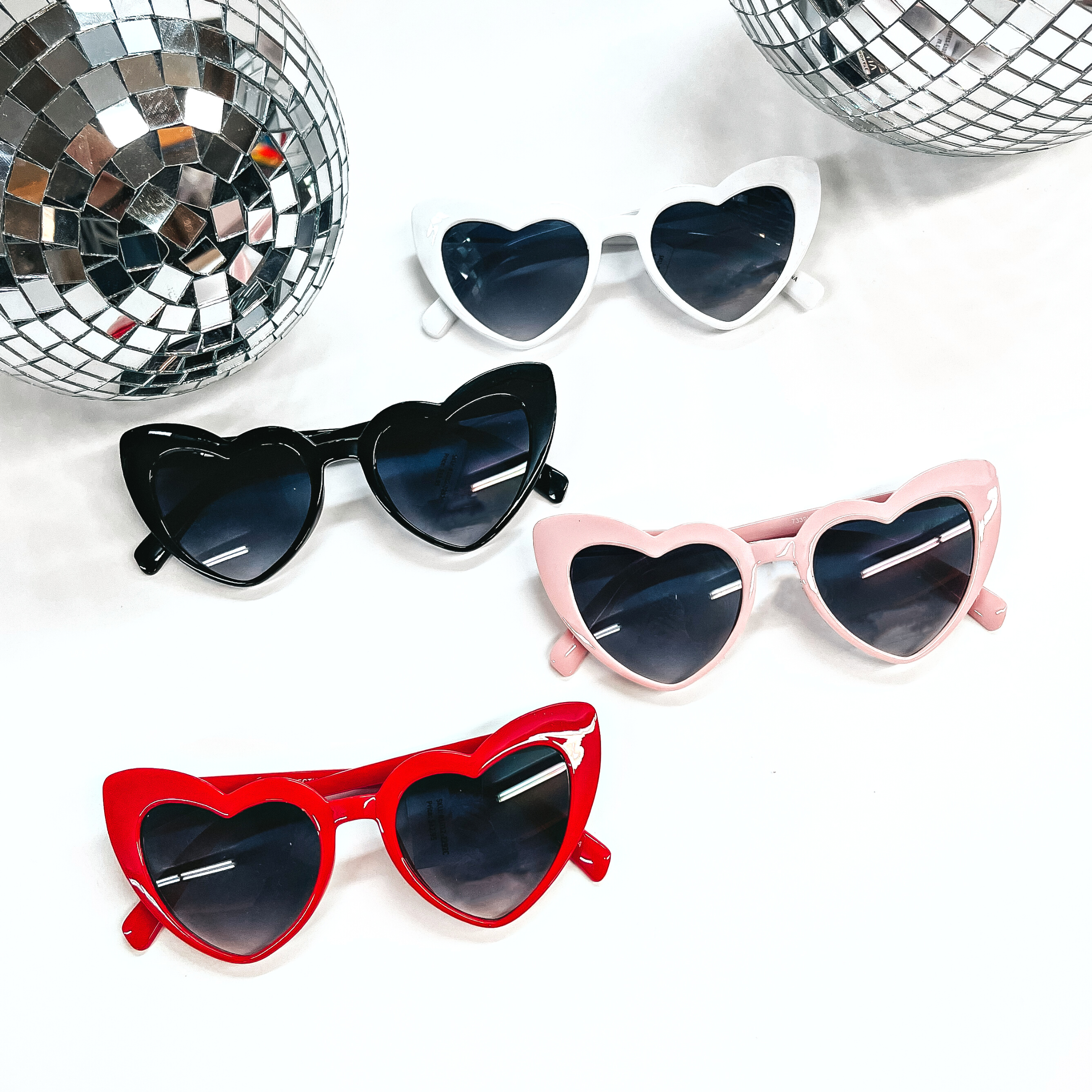 There are four heart shape sunglasses in different colors with black lenses. There are white, black, light pink, and red. All of these sunglasses are taken on a white background with two silver disco balls in the back as decor.
