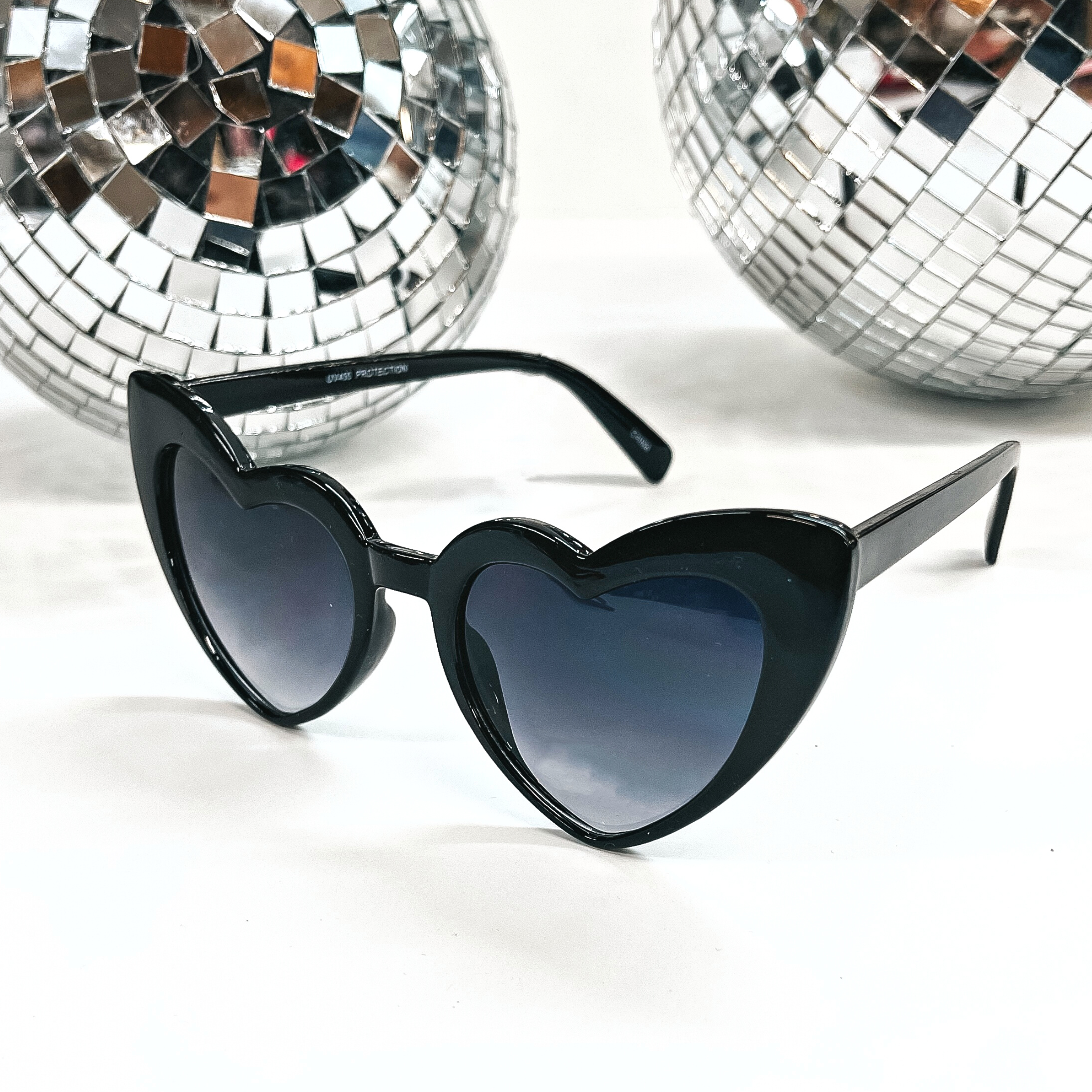 These are heart shaped sunglasses in black with black/dark grey lenses. These sunglasses are taken on a white background with two silver disco balls in the back as decor.