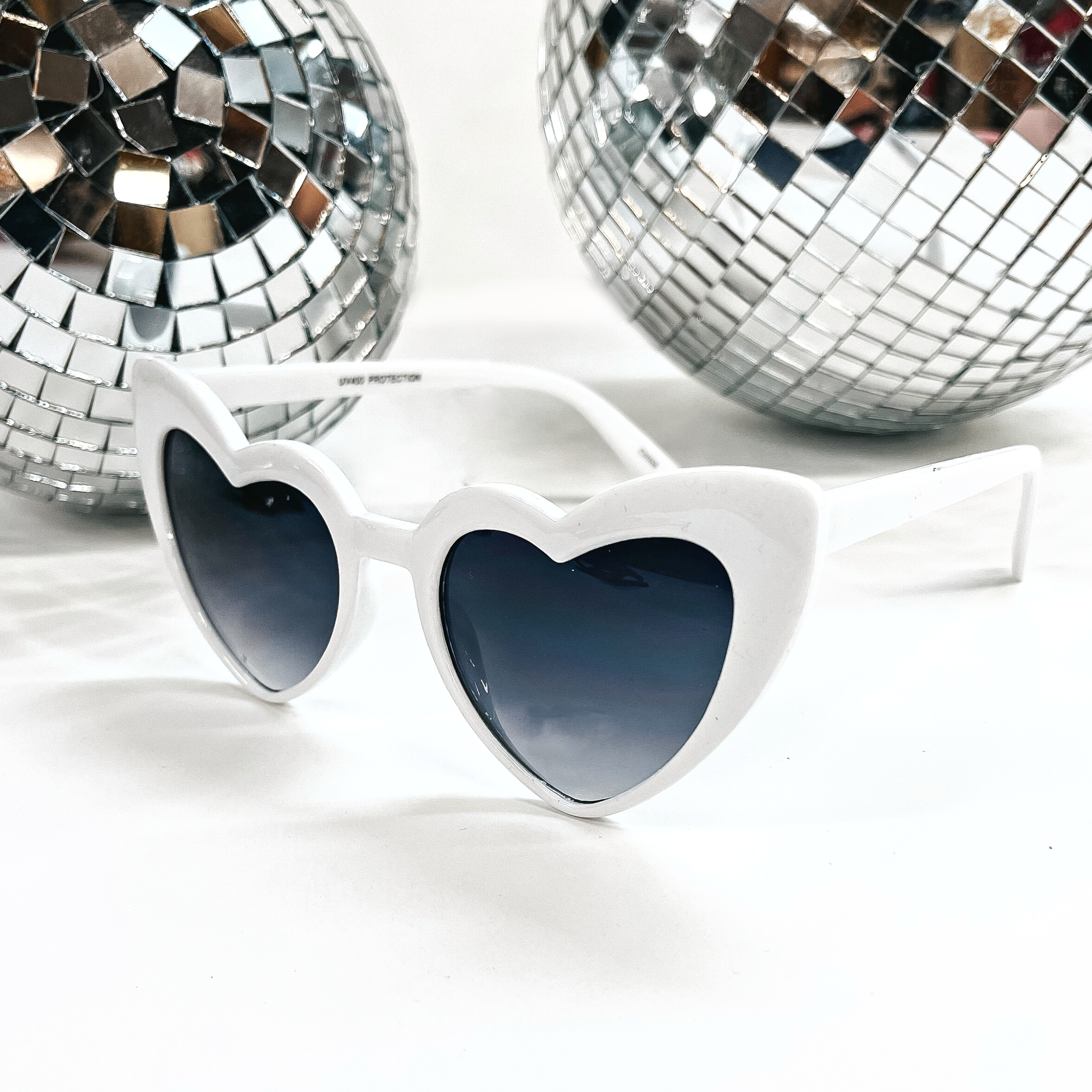 These are heart shaped white sunglasses with a black/dark grey lenses. These sunglasses are taken on a white background with two silver disco balls in the back as decor.