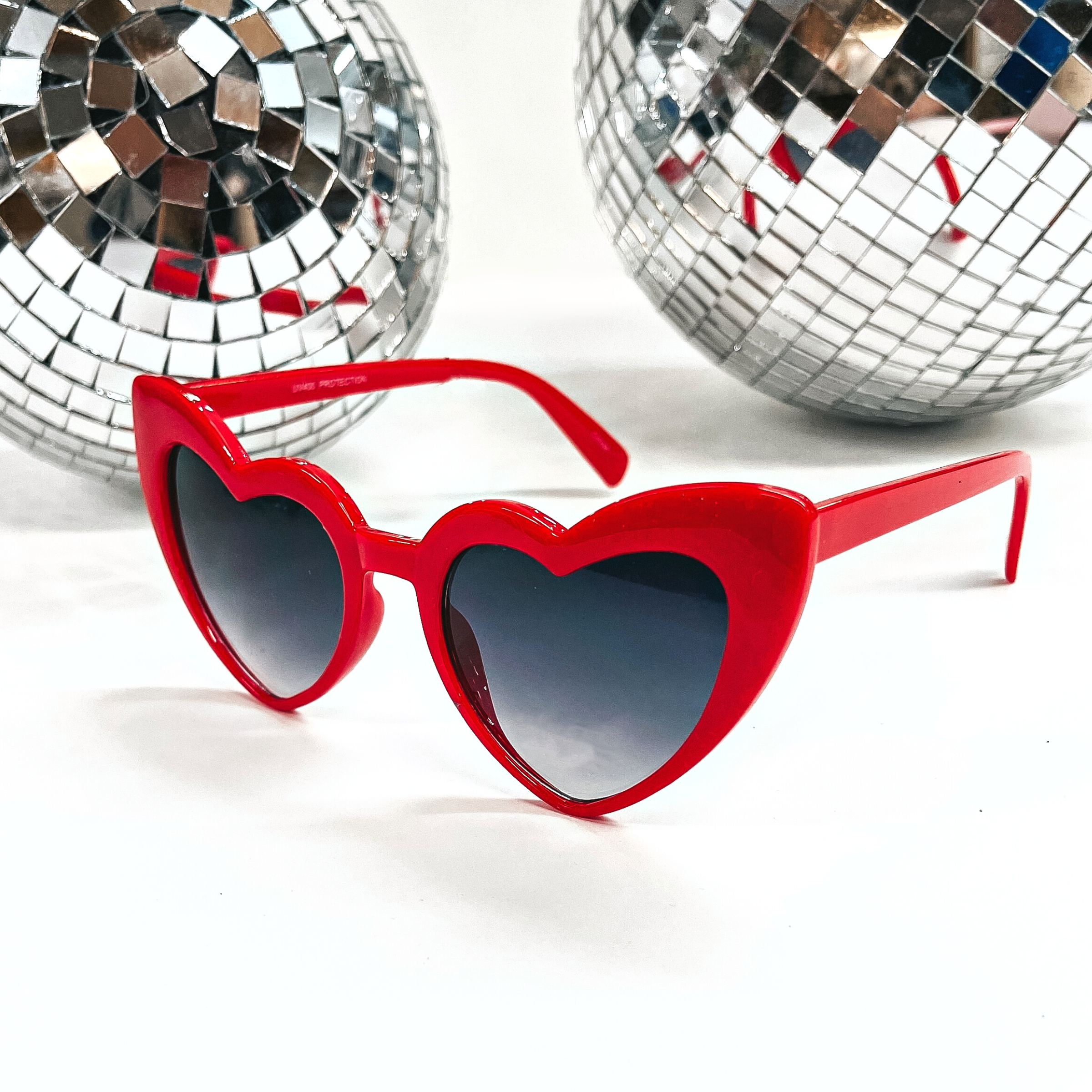 These are heart shaped sunglasses in red with a black/dark grey lenses. These sunglasses are taken on a white background with two silver disco balls in the back as decor.