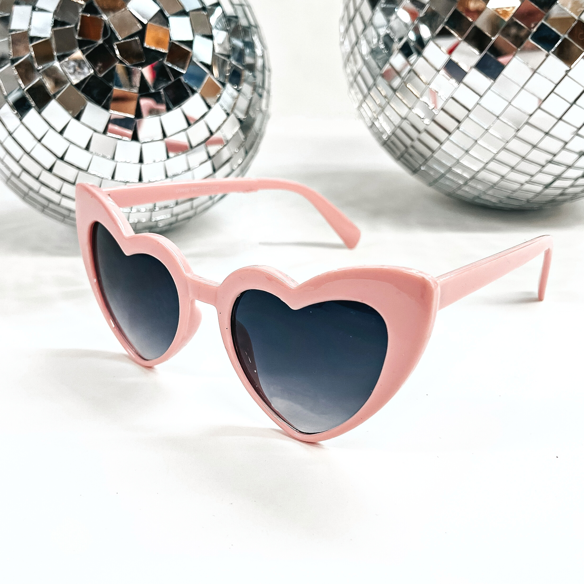 These are heart shaped sunglasses in light pink with black.dark grey lenses. These sunglasses are taken on a white background with two silver disco balls in the back as decor.