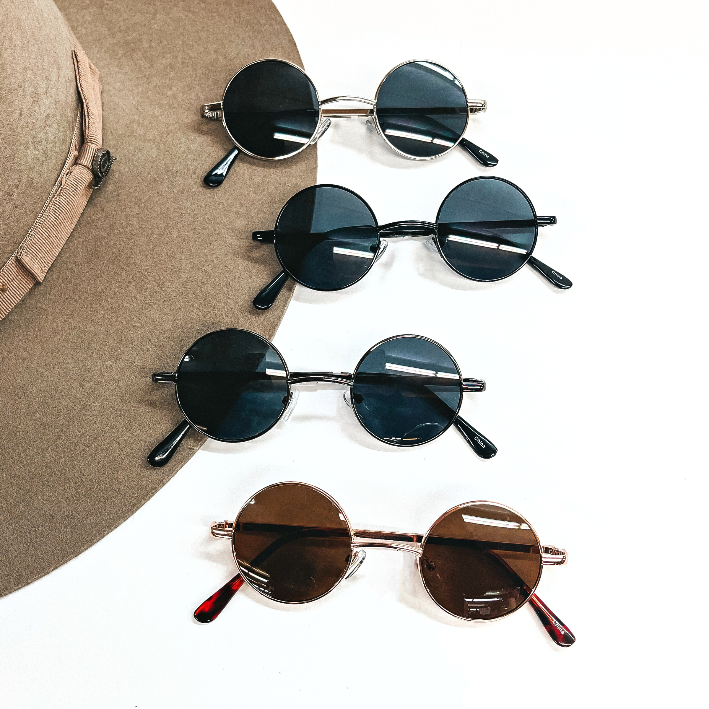 There are four pairs of sunglasses with circle lenses in different colors and frames. There are four different frame lenses in silver, black, gunmetal, and rose gold. All four pairs of sunglasses are taken on top of a white background and with a a sand felt hat in the side as decor.