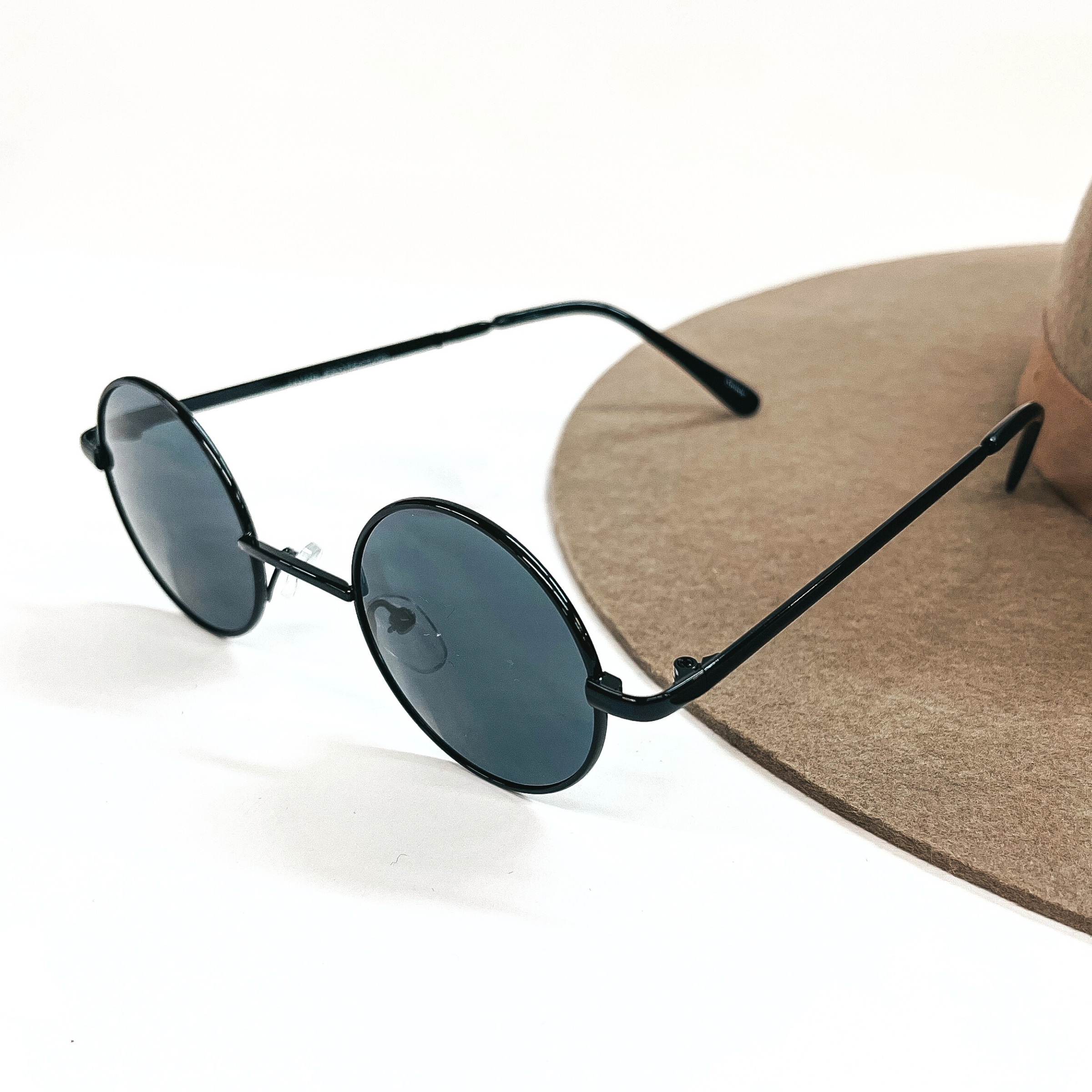 These are small circle sunglasses with black/dark grey lenses and a black outline/frame. These sunglasses are taken on a a white background and on a brown felt hat brim.