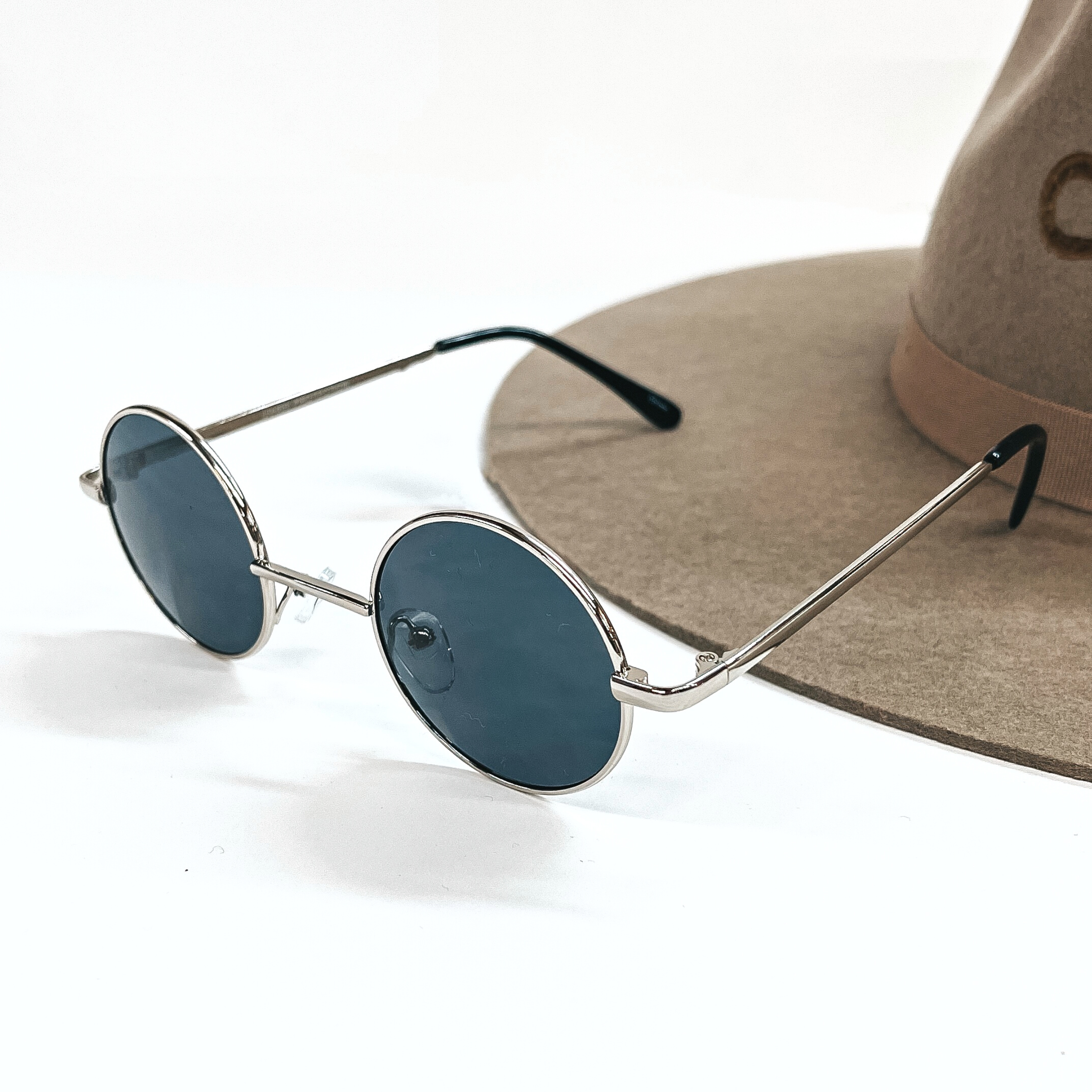 These are small circle sunglasses with dark grey lenses and a  silver outline/frame.  These sunglasses are taken on a a white background and on a brown  felt hat brim.