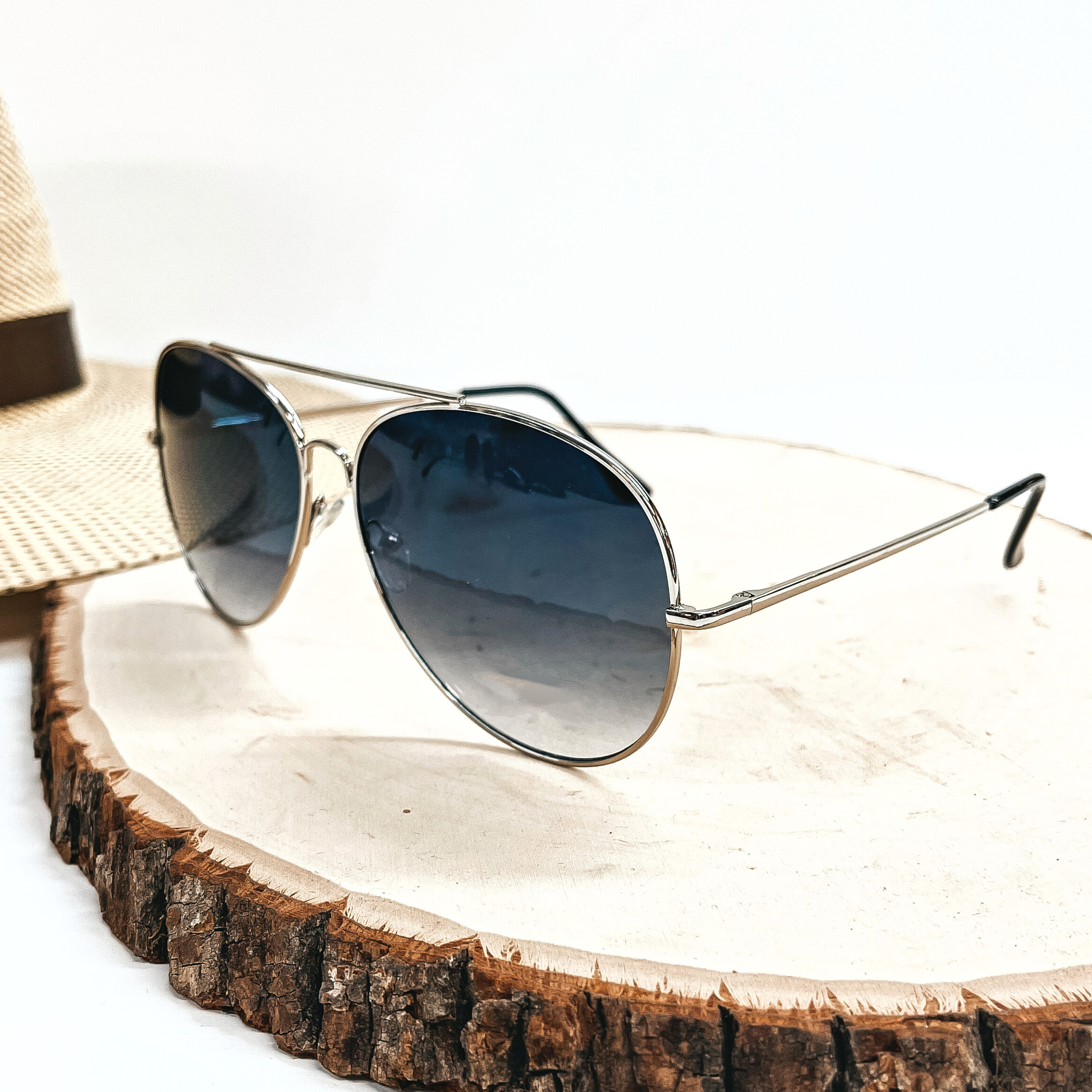 These are aviator style sunglasses with a dark grey/black lense and a silver  outline/frame. These sunglasses are taken on top of a slab of wood with a  straw hat in the back as decor.
