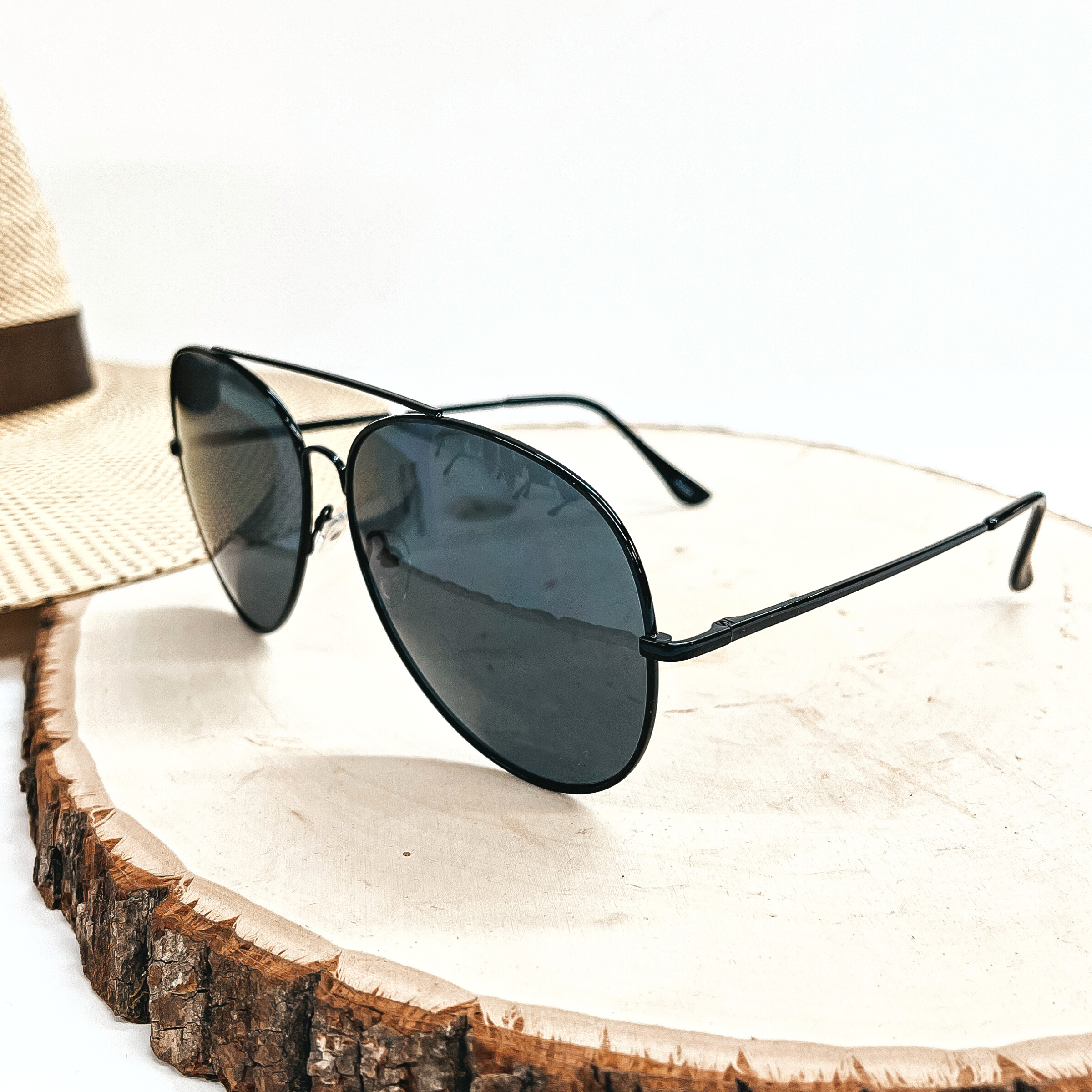 These are aviator style sunglasses with a black lense and a black  outline/frame. These sunglasses are taken on top of a slab of wood with a  straw hat in the back as decor.