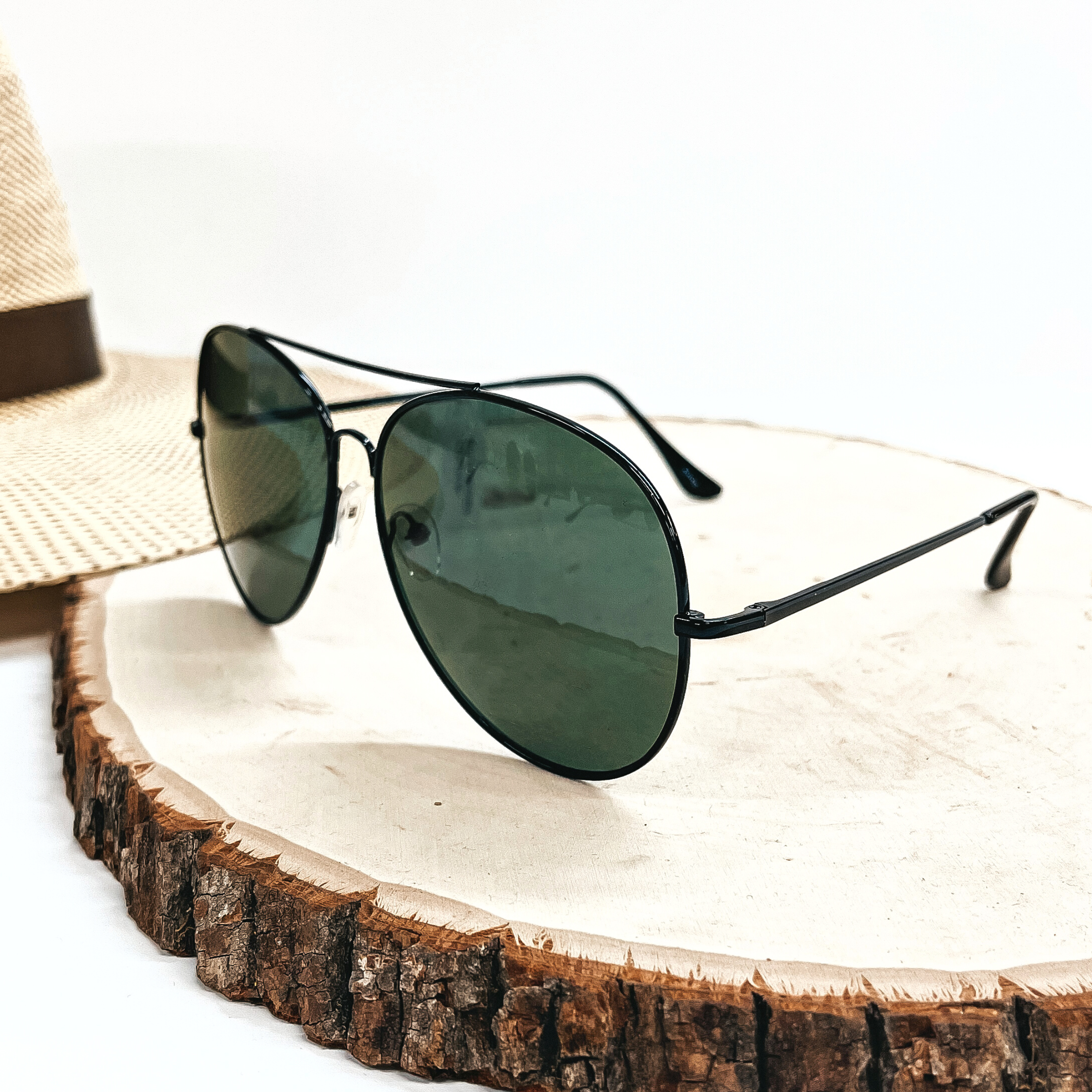 These are aviator style sunglasses with a dark green lense and a black  outline/frame. These sunglasses are taken on top of a slab of wood with a  straw hat in the back as decor.