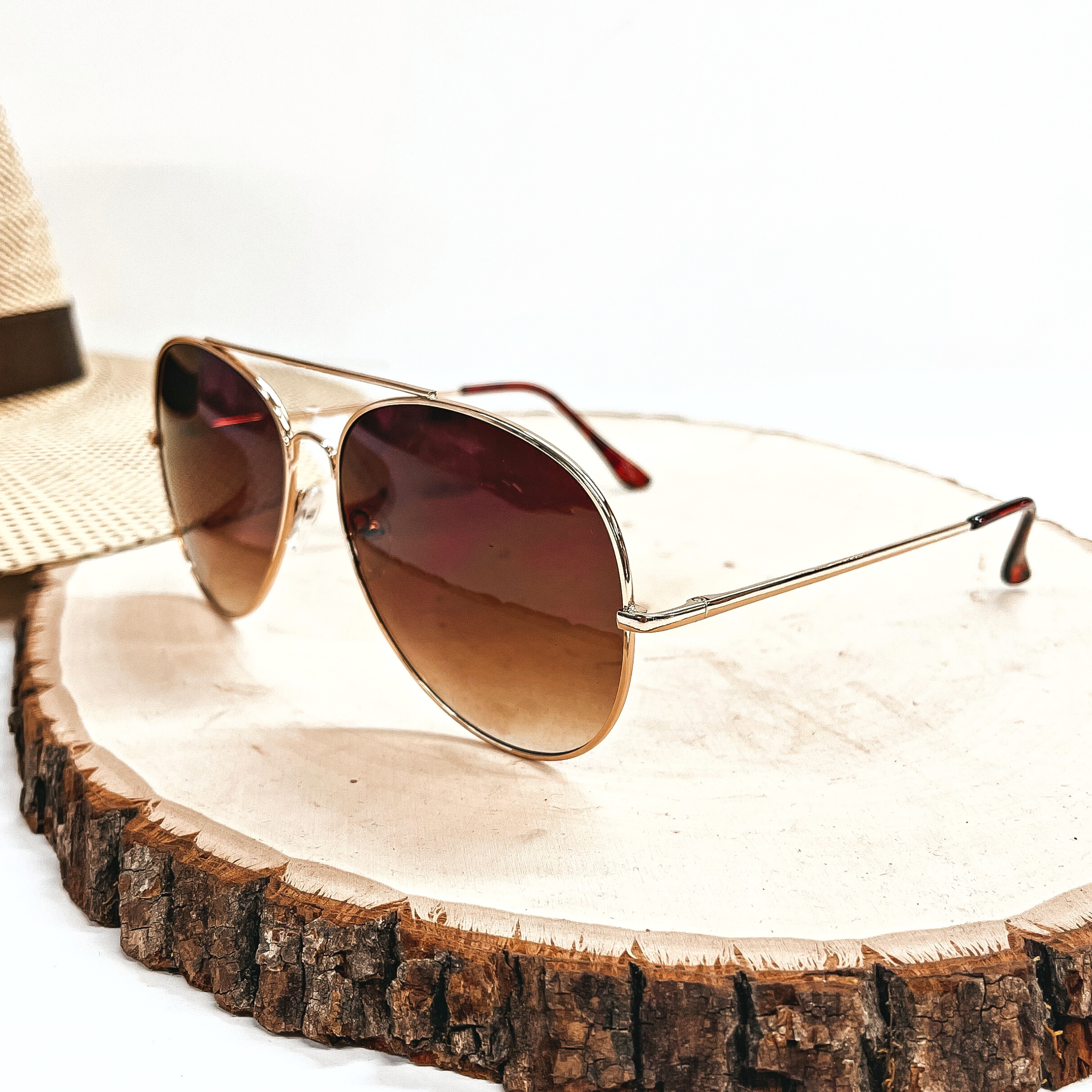 These are aviator style sunglasses with a brown lense and a gold  outline/frame. These sunglasses are taken on top of a slab of wood with a  straw hat in the back as decor.