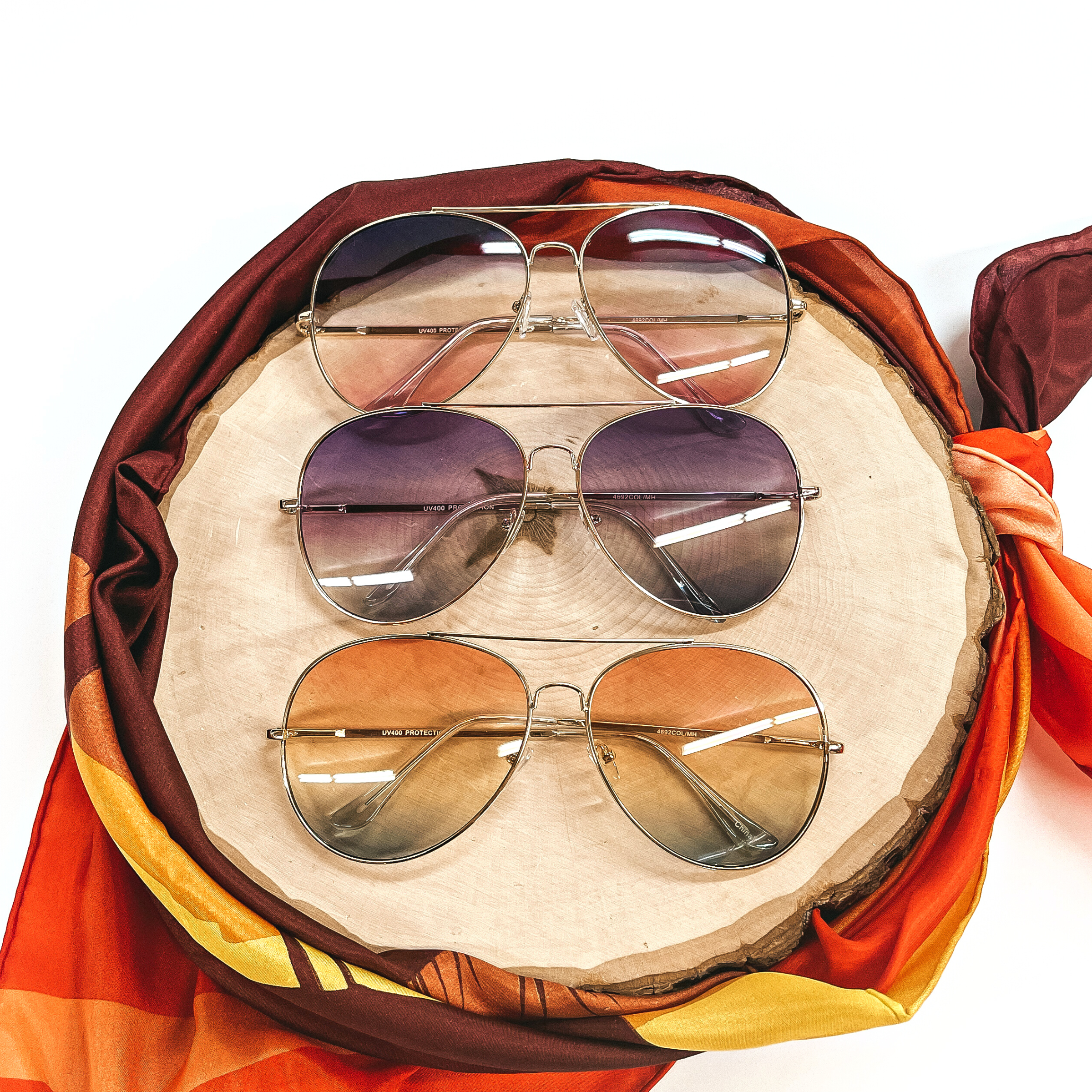 There are three pairs of aviator style sunglasses in different lense colors. From top to bottom; dark blue and pink gradient lenses, dark purple and blue lenses, and coral/orange and green lenses. All sunglasses have a gold tone outline/frame. These sunglasses are taken  on top of a wooden slate with an orange,yellow, and brown wild rag around.