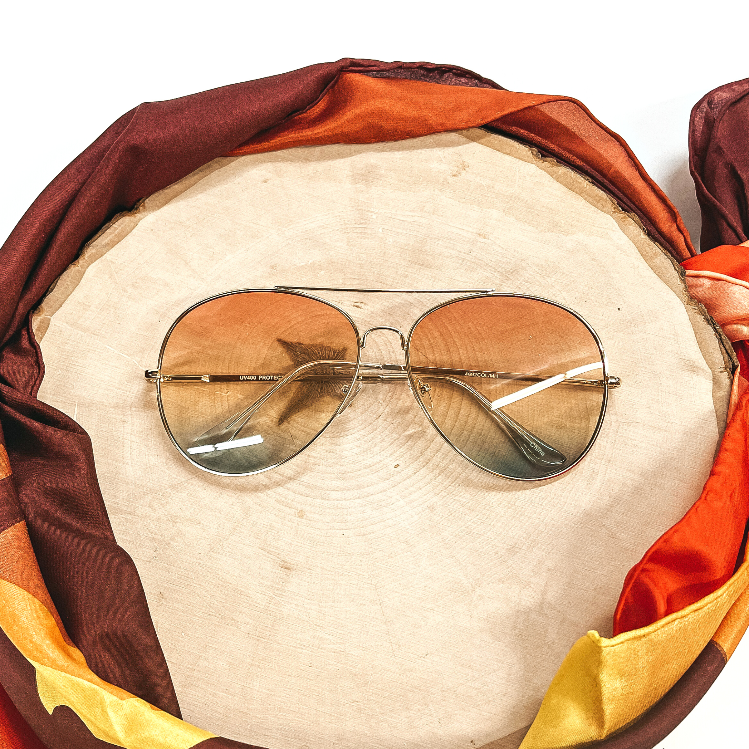 These are aviator style sunglasses with coral/orange and green gradient lenses and a gold tone outline/frame. These sunglasses are taken on top of a wooden slate and a white background. There is a brown,orange, and yellow wild rag aorund the wooden slate as decor.
