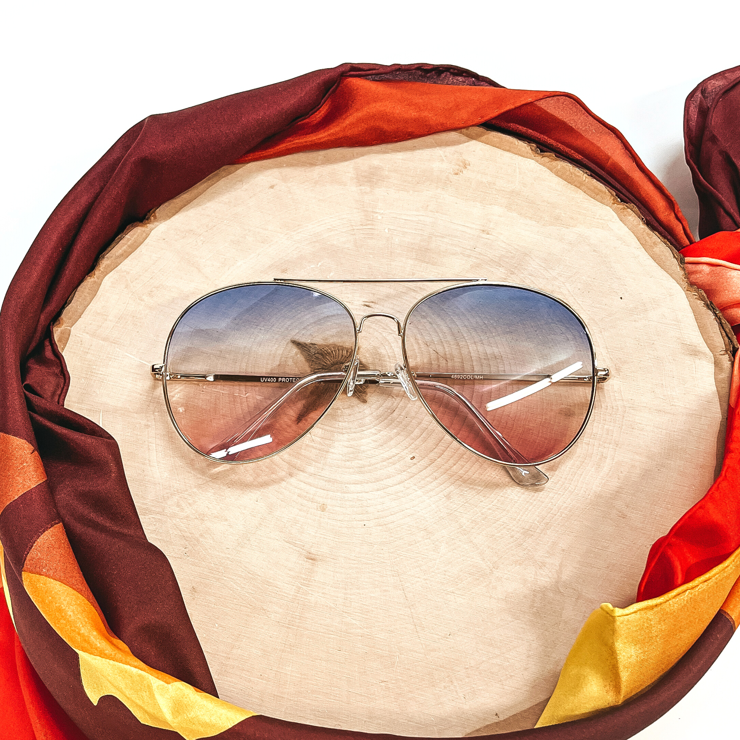 These are aviator style sunglasses with blue and light pink gradient  lenses and a gold tone outline/frame.  These sunglasses are taken on top of a wooden slate and a white background.  There is a brown,orange, and yellow wild rag around the wooden slate as decor.
