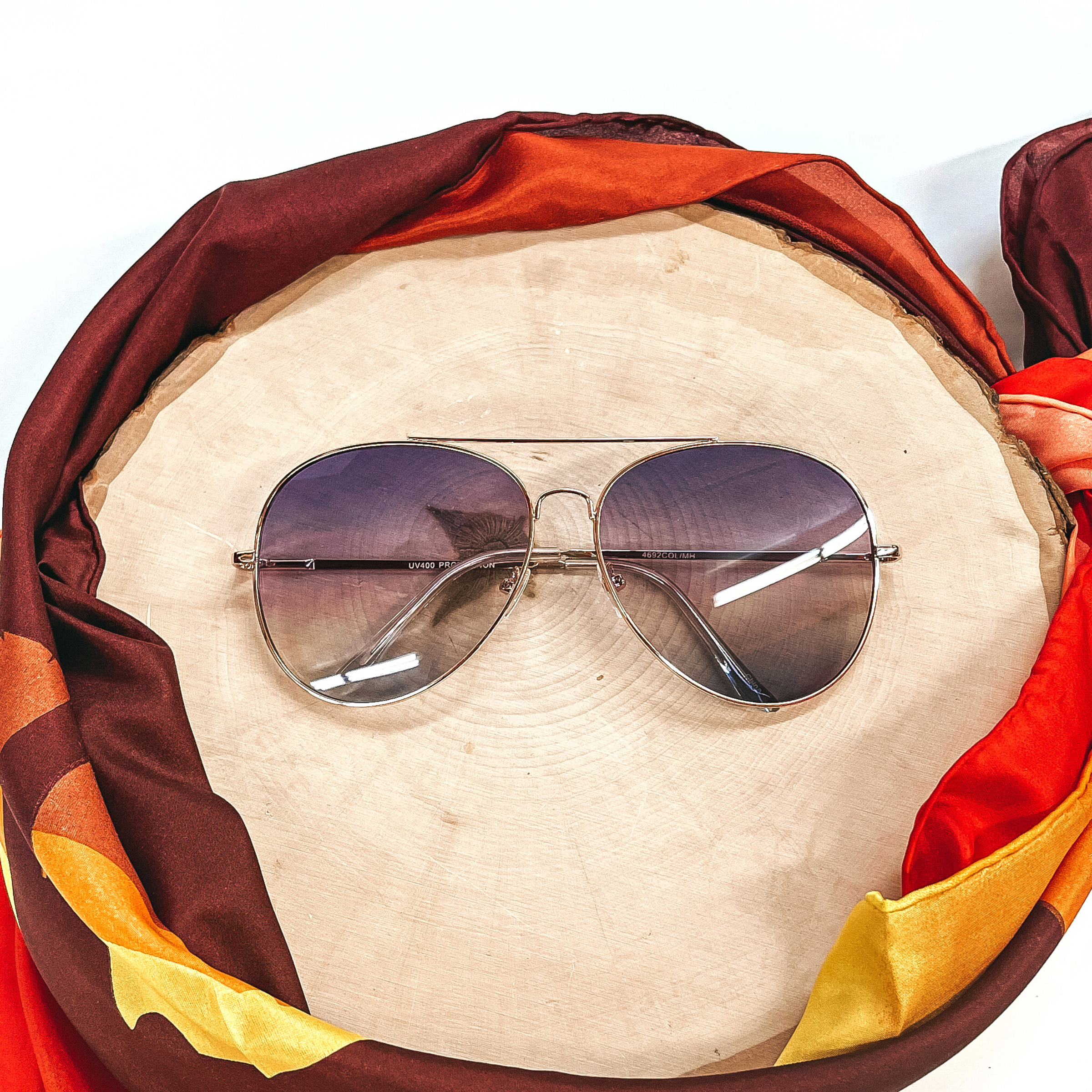 These are aviator style sunglasses with dark purple and dark blue gradient  lenses and a gold tone outline/frame.  These sunglasses are taken on top of a wooden slate and a white background.  There is a brown,orange, and yellow wild rag around the wooden slate as decor.