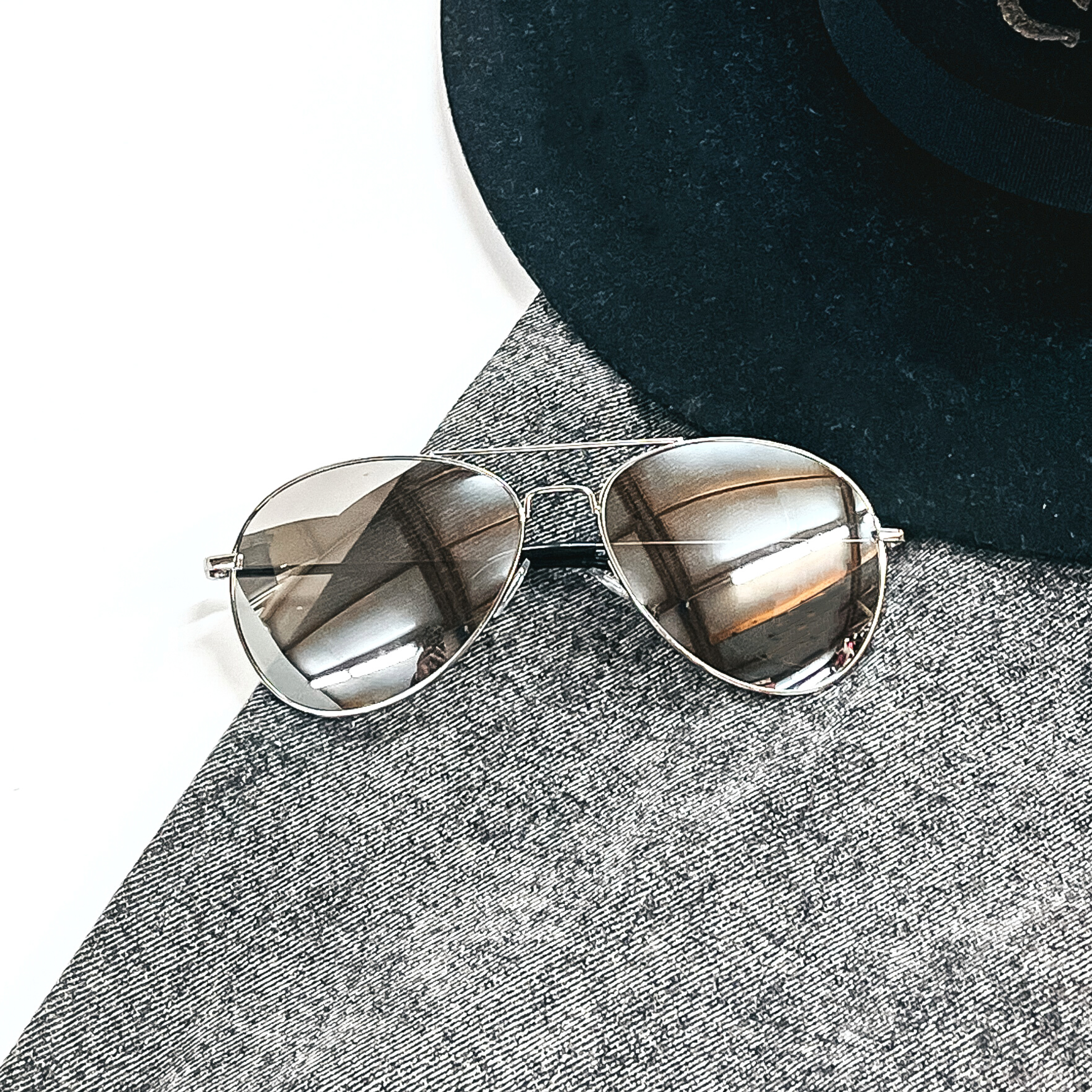 These are silver chrome mirrored sunglasses in an aviator style with a silver outline/frame. These sunglasses are taken laying on black/dak grey denim  and on a white background, with a black felt charlie horse hat in the back as decor.