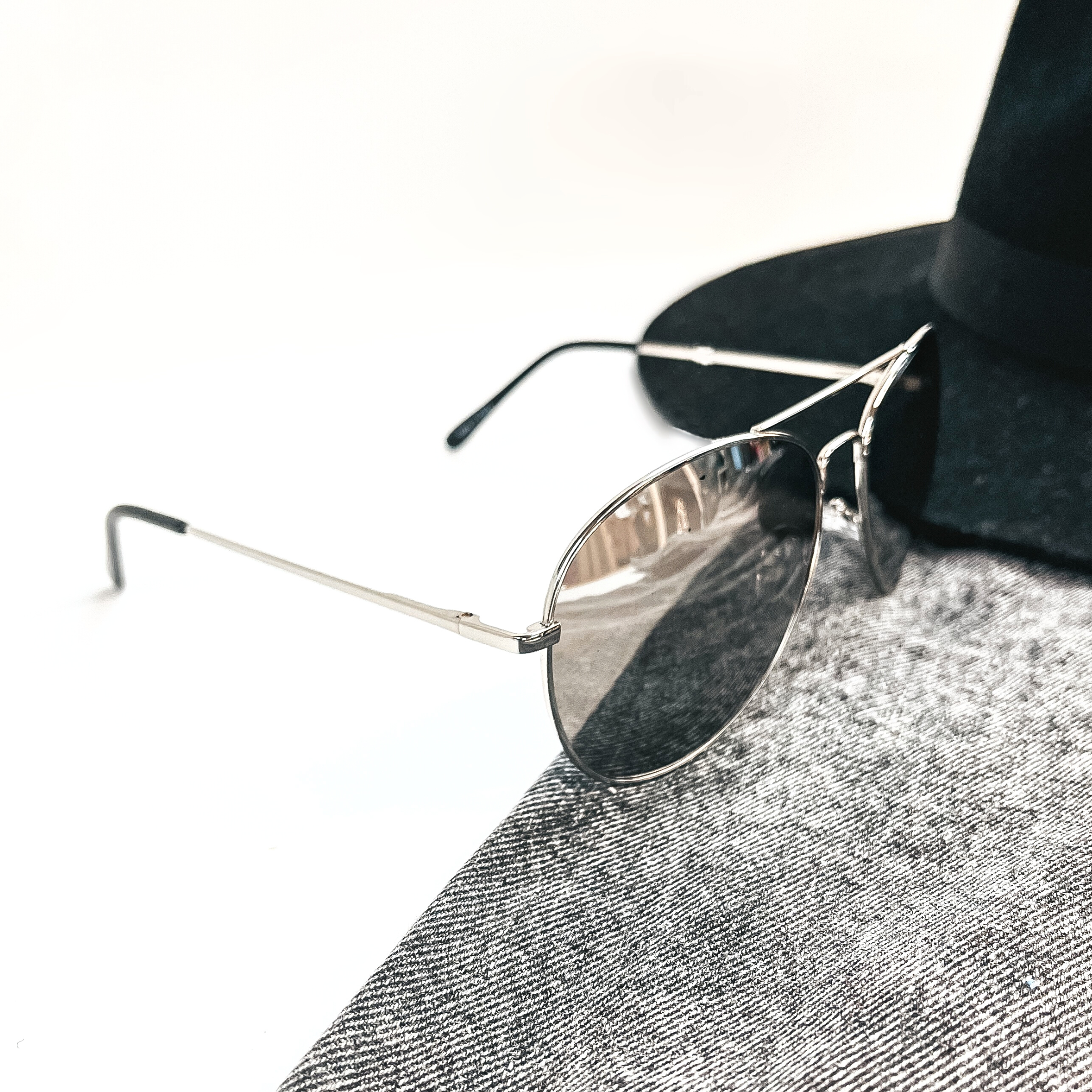 These are silver chrome mirrored sunglasses in an aviator style with silver frame/outline and black ear pieces. These sunglasses are taken on black/dark grey denim and a white background. There is a black felt hat in the back as decor.