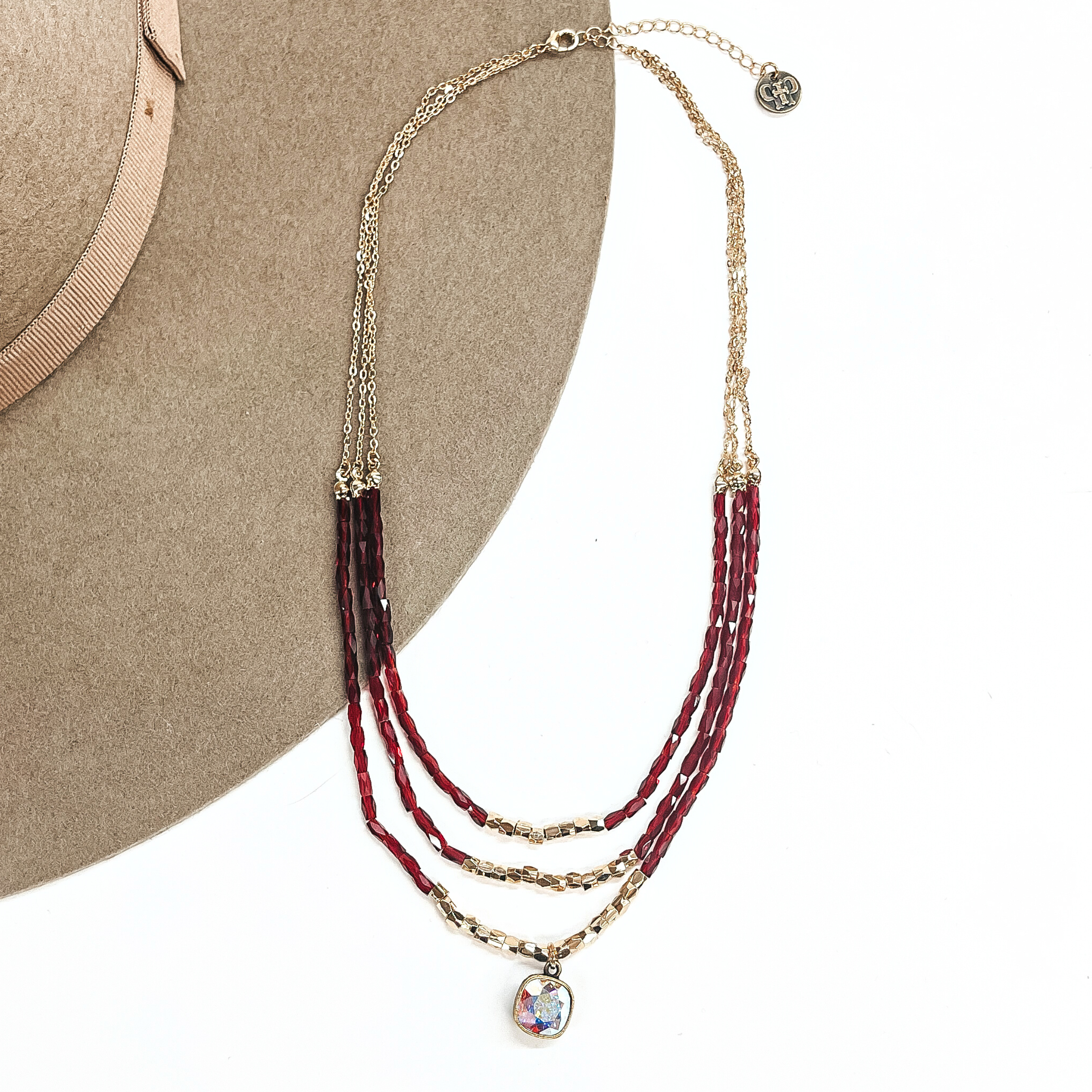 This is a three strand gold necklace with long burgundy beads in the bottom with gold tone beads in the bottom center of each strand. The longest strand has an ab cushion cut crystal in a gold setting. This necklace is taken on a light brown felt hat and on a white background.