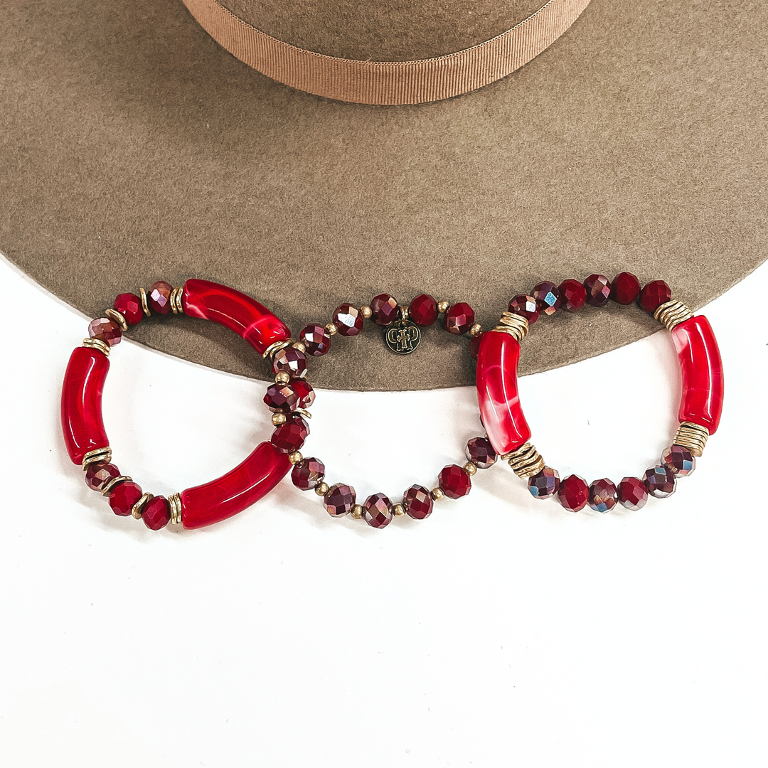 These are a set of three bracelets, two have tubes with crystals and thhe middle one has crystals all around, From left to right; three red tubes and three wine colored crystals beads in between each tube with gold spacers. The middle one has wine colored crystal beads with gold spacers. The right one has two marble red/white tubes with gold spacers and six wine colored crystal beads on each sides. These three bracelets are taken on a white background and on a light brown felt hat brim.