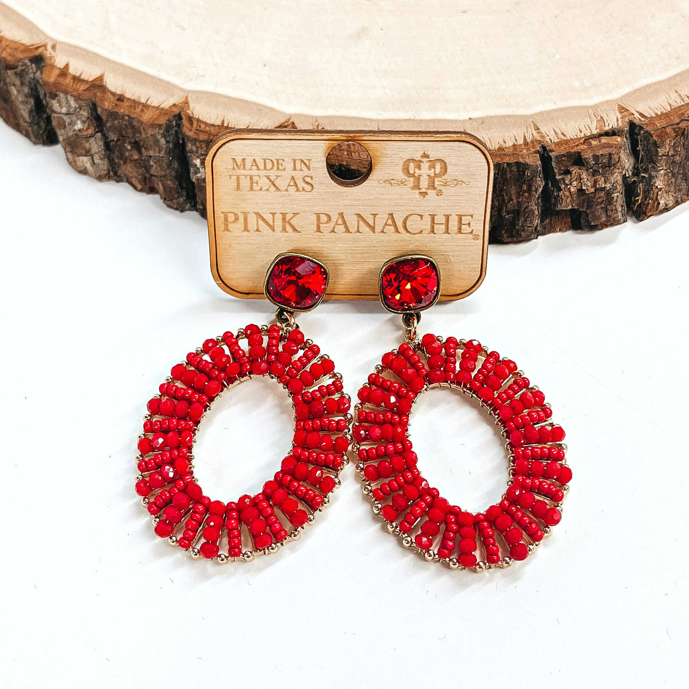 These are red cushion cut post back earrings with an oval drop. The earrings are in a gold setting with small red beads all around the oval. These earrings are taken on a Pink Panache earrings holder. These earrings are taken leaning up against a slab of wood and on a white background.