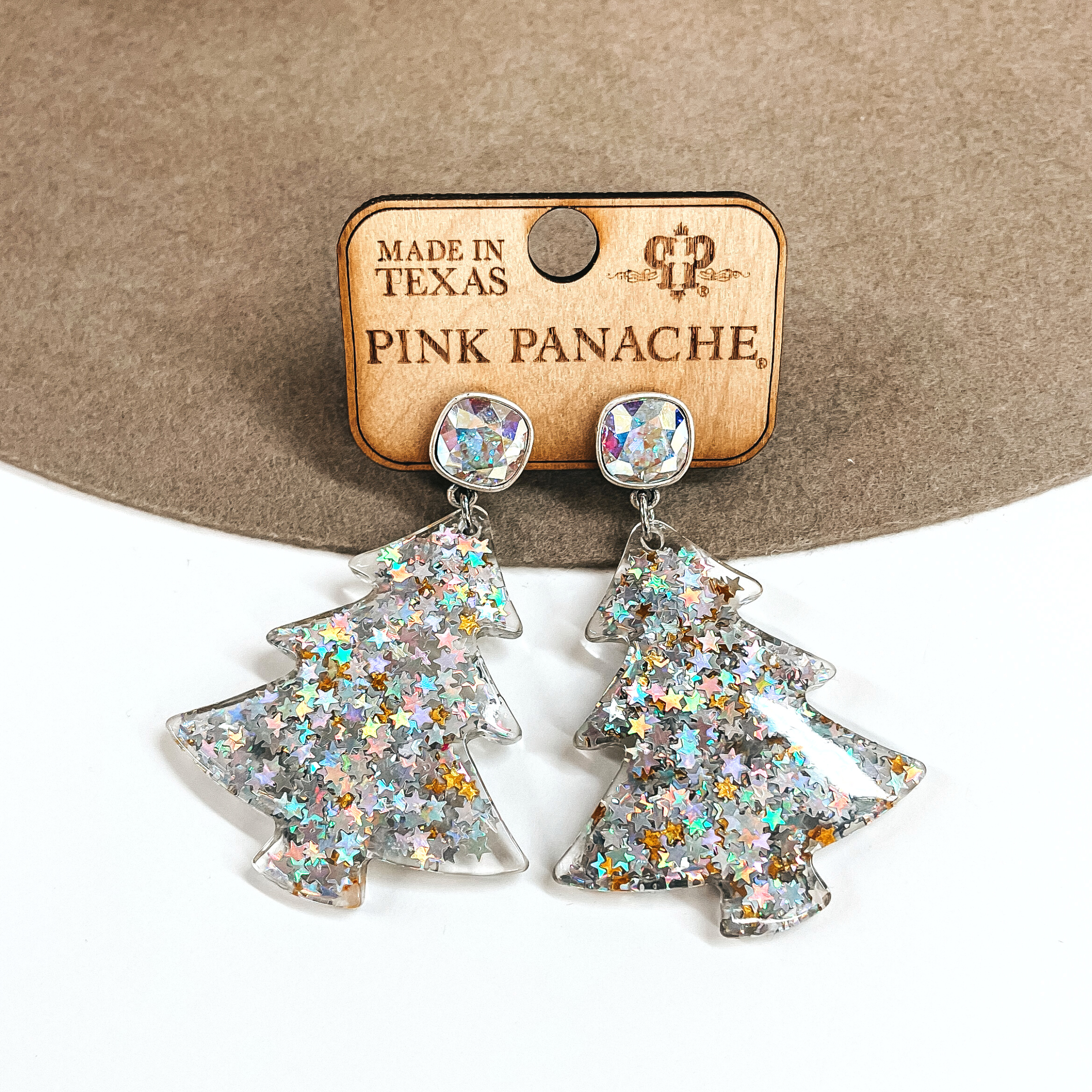 These are ab cushion cut crystal post earrings with an acrylic christmas tree drop. The christmas tree has silver and holographic glitter in small stars. These earrings are placed on a Pink Panache earrings holder. These earrings are taken on a brown felt hat brim and on a white background.