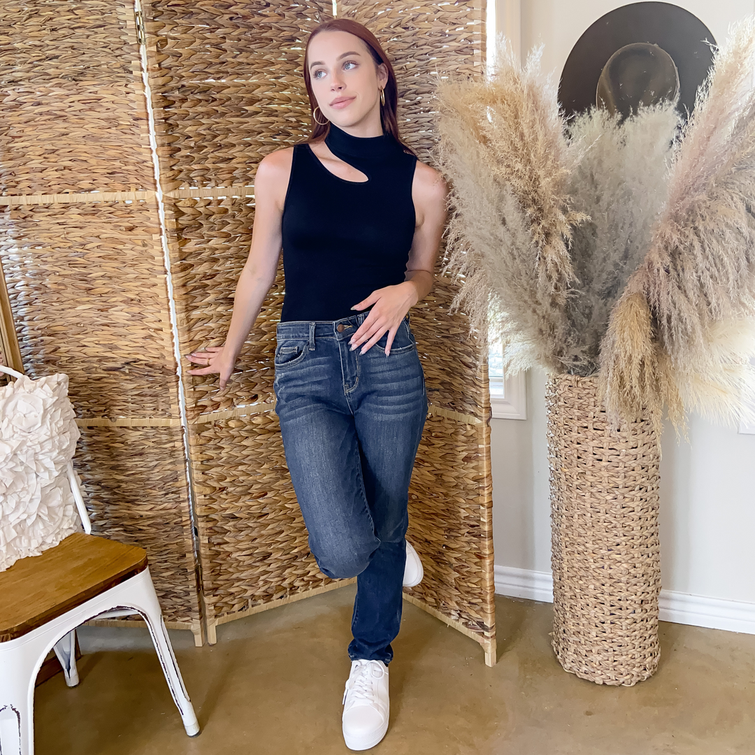 Model is wearing a black, cutout, mock neck ribbed top. She is also wearing gold hoop earrings, dark wash jeans, and white sneakers.