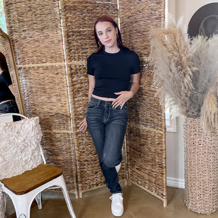 Model is wearing a black, high neck cropped short sleeve top with dark blue jeans. She is also wearing gold hoops and white tennis shoes.