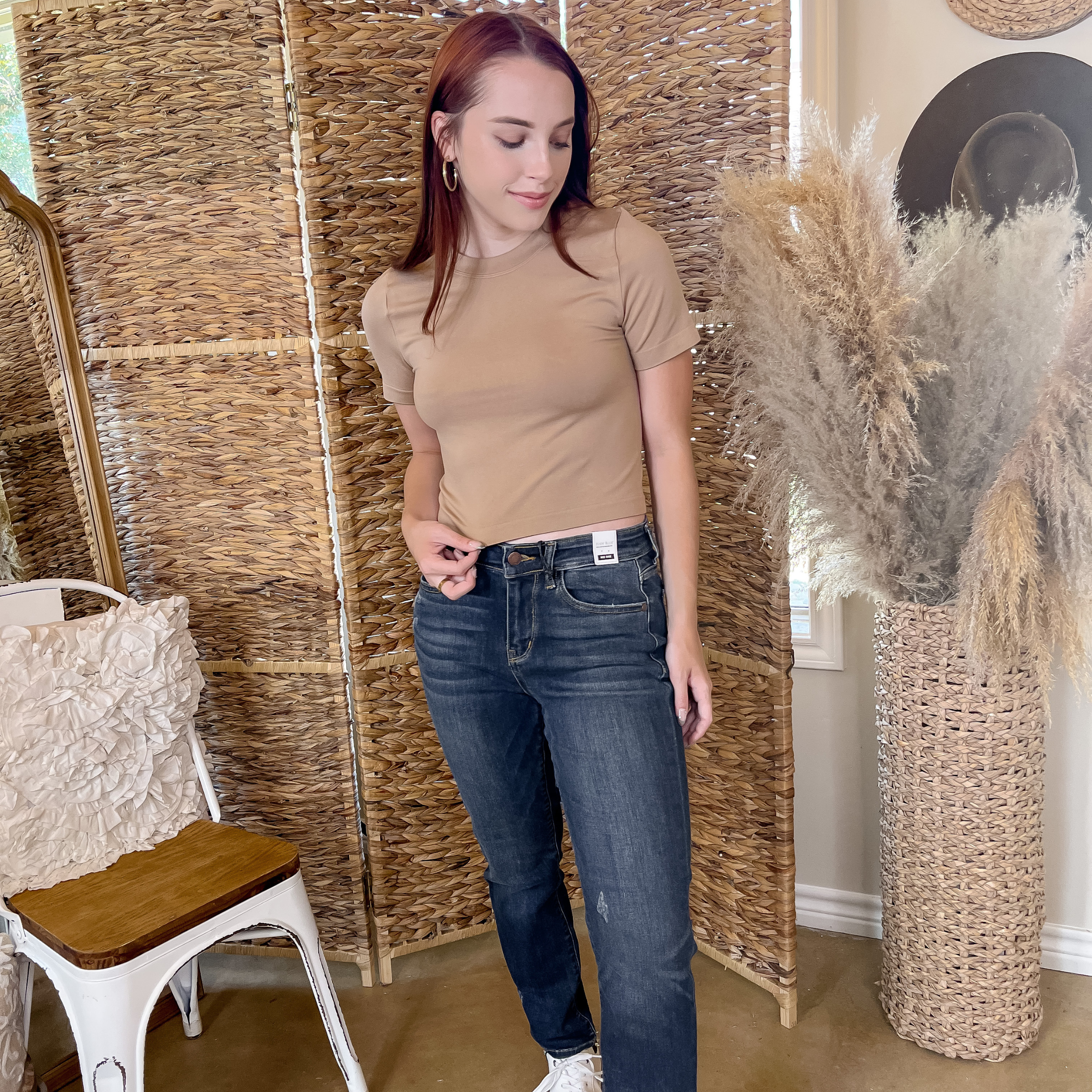 Model is wearing a nude, high neck cropped short sleeve top with dark blue jeans. She is also wearing gold hoops and white tennis shoes.