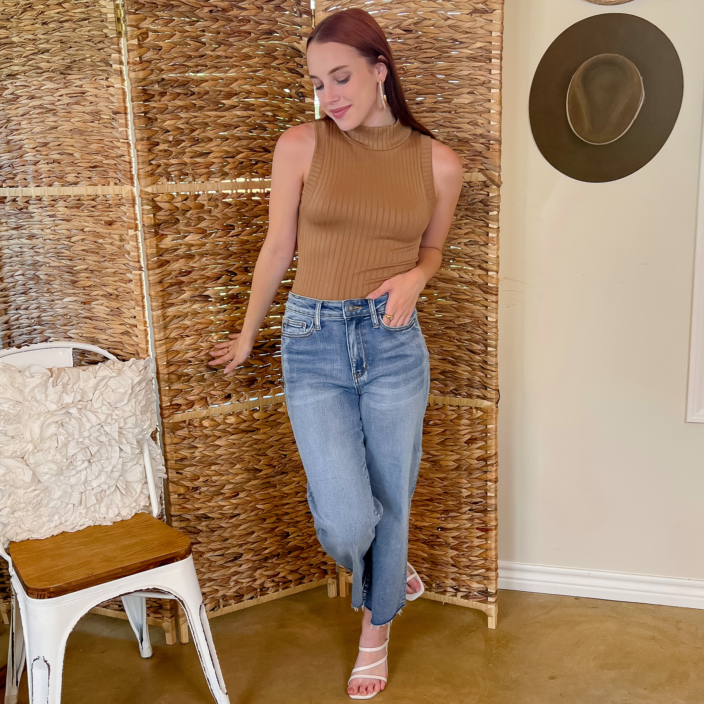 Model is wearing a high neck, ribbed tank top in mocha/brown.  She is also wearing a long gold earrings, light wash jeans, and white strappy  heels.