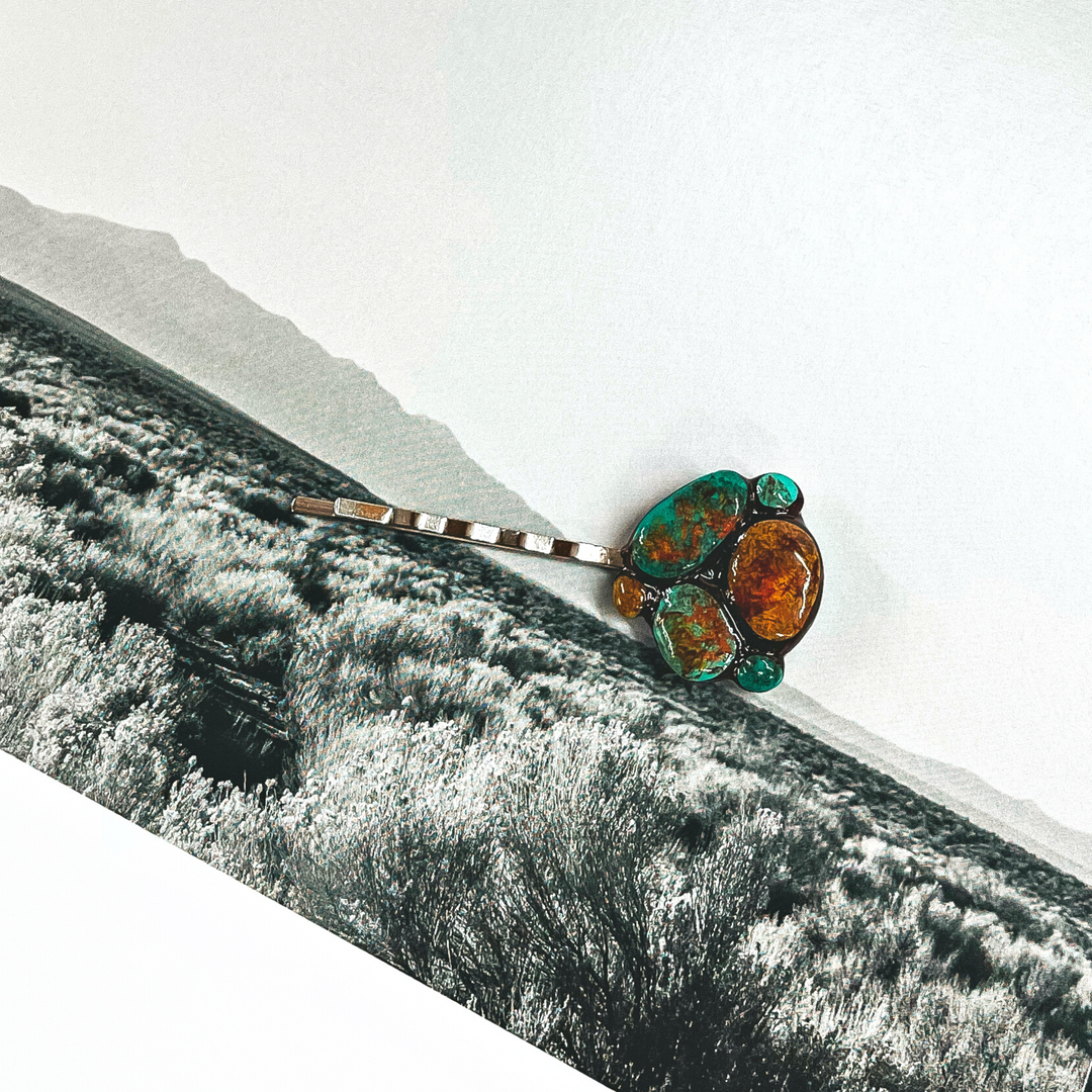 This is a silver tone bobby pin with a wanderlust clay pendant on the end of the pin. The wanderlust clay piece is turquoise/teal and brown. This bobby pin is laying on a black and white outdoor book page.